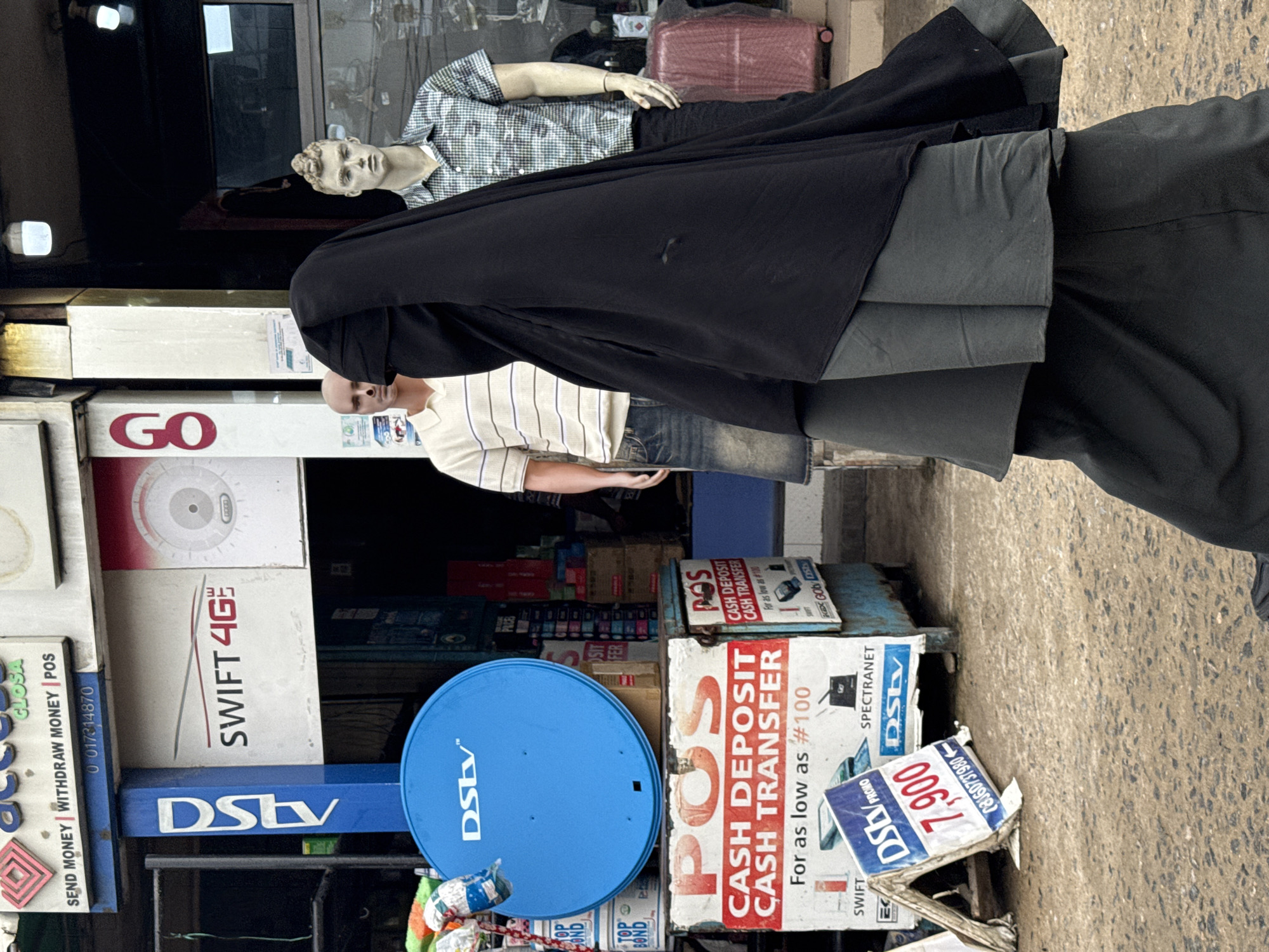 The image depicts a street scene in Lagos, Nigeria, with a store facade on the left. The store is advertising DStv and Swift 4G, featuring a blue DStv satellite dish and signs promoting cash deposit/transfer services. The signage indicates prices and phone numbers. In the foreground, a woman is seen wearing a black hijab. Inside the store, mannequins are partially visible, one with a checkered shirt and another with a striped shirt. The background displays various products and boxes within the store. The street surface is visible on the right.