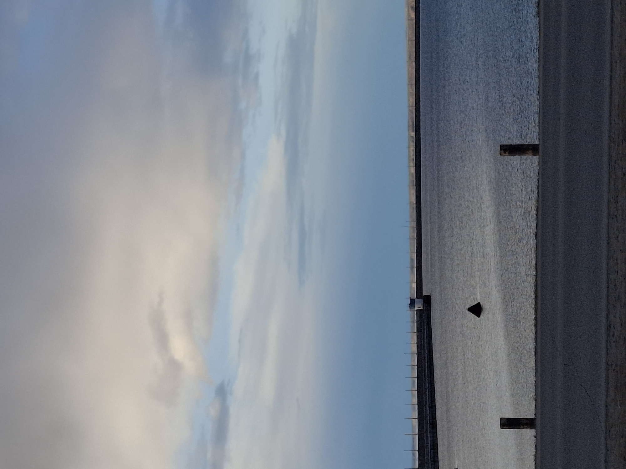 The image presents a serene landscape featuring a vast body of water under a partially cloudy sky. The scene is oriented horizontally, with the sky occupying the upper portion and the water filling the mid-ground, bordered by an embankment or road at the bottom.
The sky exhibits a blend of light blue patches and soft, diffused white and grey clouds, suggesting either an overcast day or a time of day with soft, ambient light. Below the horizon, a large expanse of water, likely a lake or reservoir, stretches into the distance. Its surface is textured with subtle ripples or reflections.
In the foreground of the water, several dark, rectangular posts protrude vertically from the surface. A distinct small, dark triangular object floats in the water further out. In the far distance, a faint line of smaller posts or a long, low structure extends horizontally across the water, possibly indicating a dam, a bridge, or a distant shoreline feature. A small, light-colored square object is also discernible on the distant left horizon at the water's edge.
The bottom edge of the image reveals a wide, textured, dark-toned surface, which appears to be a paved road, an embankment, or a wide shore running alongside the water. The scene is static, with no visible human activity or discernible text. The overall atmosphere is one of tranquility and openness. The location context indicates this scene is in Chaikovsky, Russia, likely showcasing a view of the Kama Reservoir.