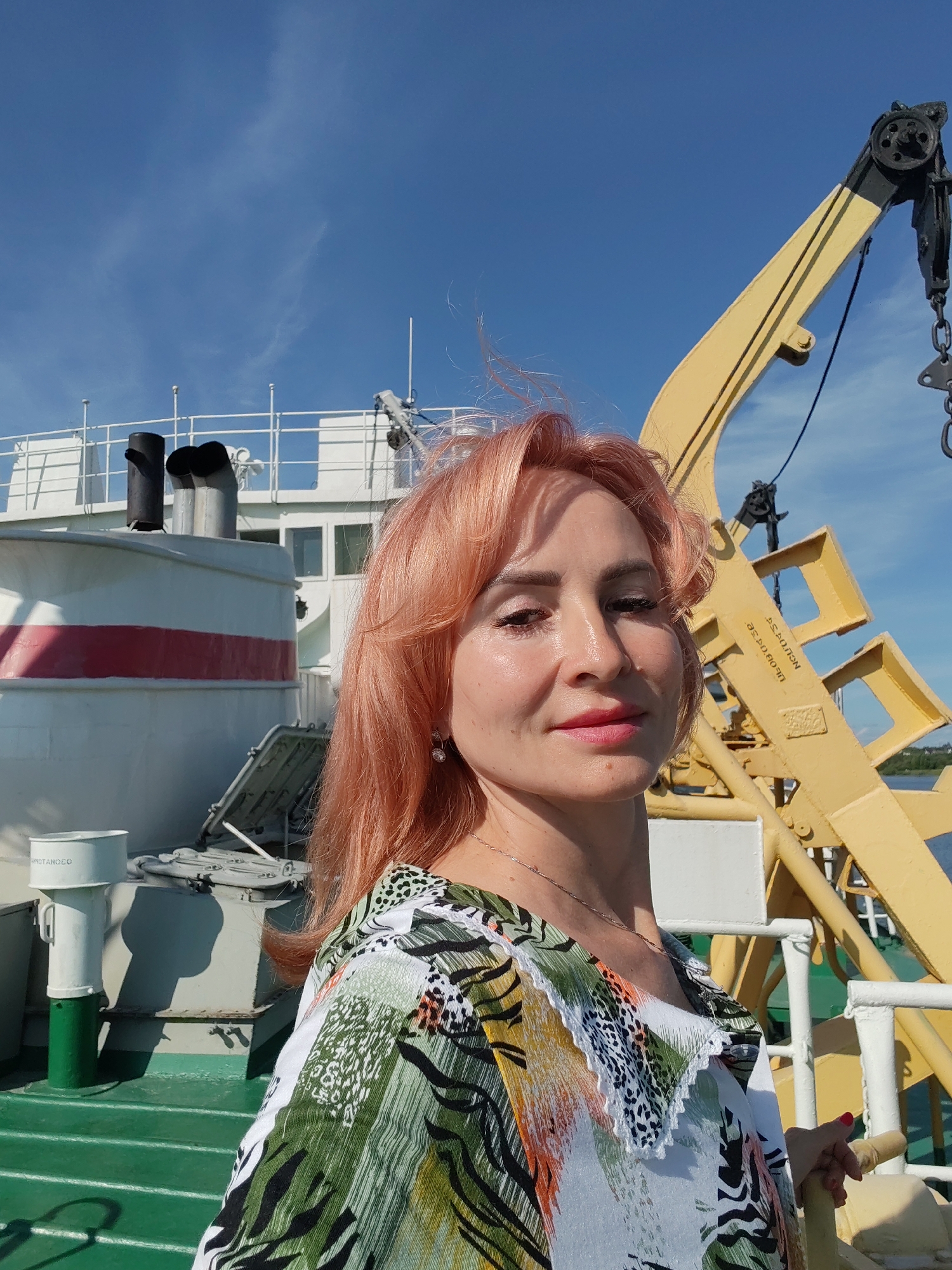 This is a medium shot selfie of a woman with reddish-pink hair and fair skin, taken outdoors on a sunny day. She is looking towards the camera with a slight smile. She is wearing a colorful top with a white, green, black, and orange abstract print and a delicate silver necklace. Her hair is styled with gentle waves, and she has subtle makeup on. In the background, a large white ship with a red stripe is visible, along with yellow crane equipment and a clear blue sky with wispy clouds. The scene appears to be on the deck of a ship, with a green deck surface in the foreground. The overall mood is bright and cheerful. There is visible text on the yellow crane which reads "ASDN09080" and "SAYMON". The location is identified as Cherepovets, Russia.