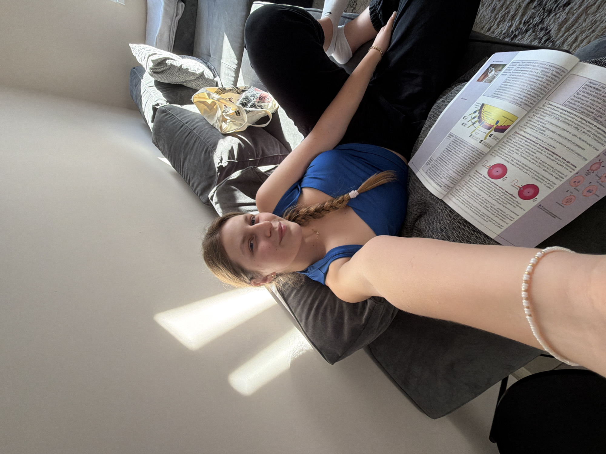A young woman is lying on a gray sofa. She is wearing a blue top and black pants, and has a braid in her hair. She is holding a book open, with diagrams and text visible on the pages. Beside her are some pillows and a tote bag. The scene is lit by sunlight, which casts shadows on the wall. No location information is available.
