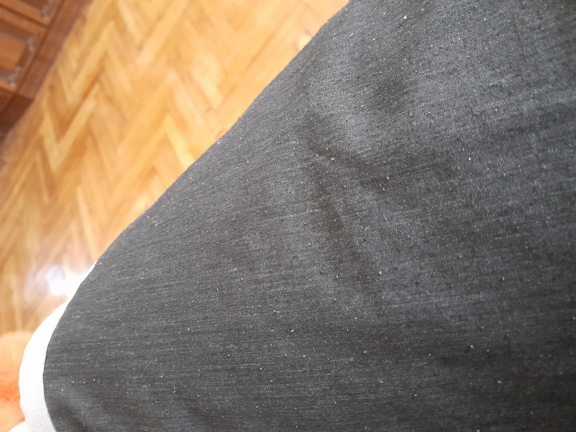 The image shows a close-up view of a person's dark gray fabric clothing, possibly jeans or trousers, with small white specks of lint or dust visible on the surface. The fabric is creased in several areas, indicating it is being worn. In the background, a polished wooden floor with a herringbone pattern is visible, along with what appears to be the base of wooden furniture. The lighting suggests an indoor environment. There are no identifiable landmarks or features that would indicate a specific location in Yerevan, Armenia.
