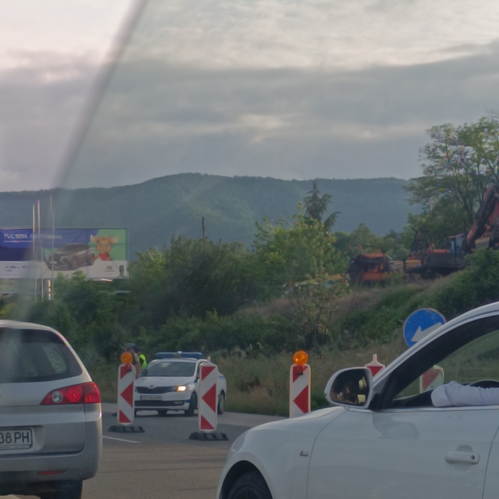 The image depicts a scene in Dolno Varshilo, Bulgaria, possibly near a highway construction or traffic control area. In the foreground, the rear of a silver car is visible on the left, with a license plate reading "38 PH". To the right, the front portion of a white car is seen, with a side mirror reflecting a distorted view.
In the middle ground, a police checkpoint is set up. A white police car with flashing blue lights is positioned behind two red and white striped traffic cones, each topped with an orange flashing light. A police officer, wearing a high-visibility vest over a uniform, is standing near the police car, interacting with someone. A blue and white directional sign is also present in this area.
Further in the background, several construction vehicles are visible, including what appears to be an excavator. The landscape features lush green trees and rolling hills under a cloudy sky. A billboard with advertisements for "Tucson GO" and a cartoon character is also visible on the left side of the image, suggesting the location is near a main road.