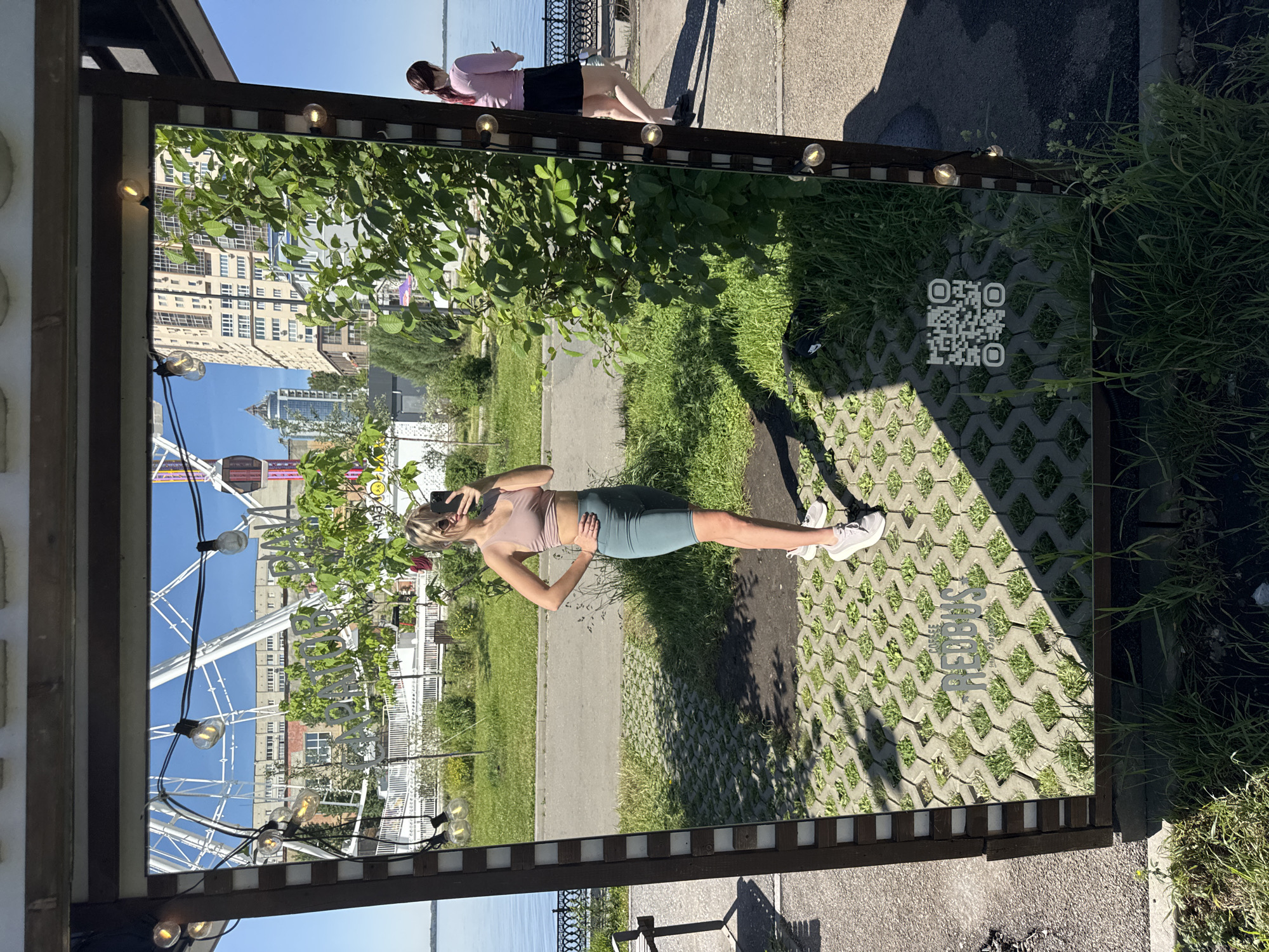 The image shows an outdoor scene in Saratov, Russia. A woman in athletic wear (tank top, shorts, and sneakers) is taking a selfie in front of a mirrored structure with decorative lighting. The mirror reflects a Ferris wheel, waterfront, buildings, and foliage. To the right of the mirrored structure, there's a grassy area and a brick-paved surface with the word "BEDBUS" printed on it. Another woman stands in the background alongside a metal fence, facing the water. The day appears sunny.