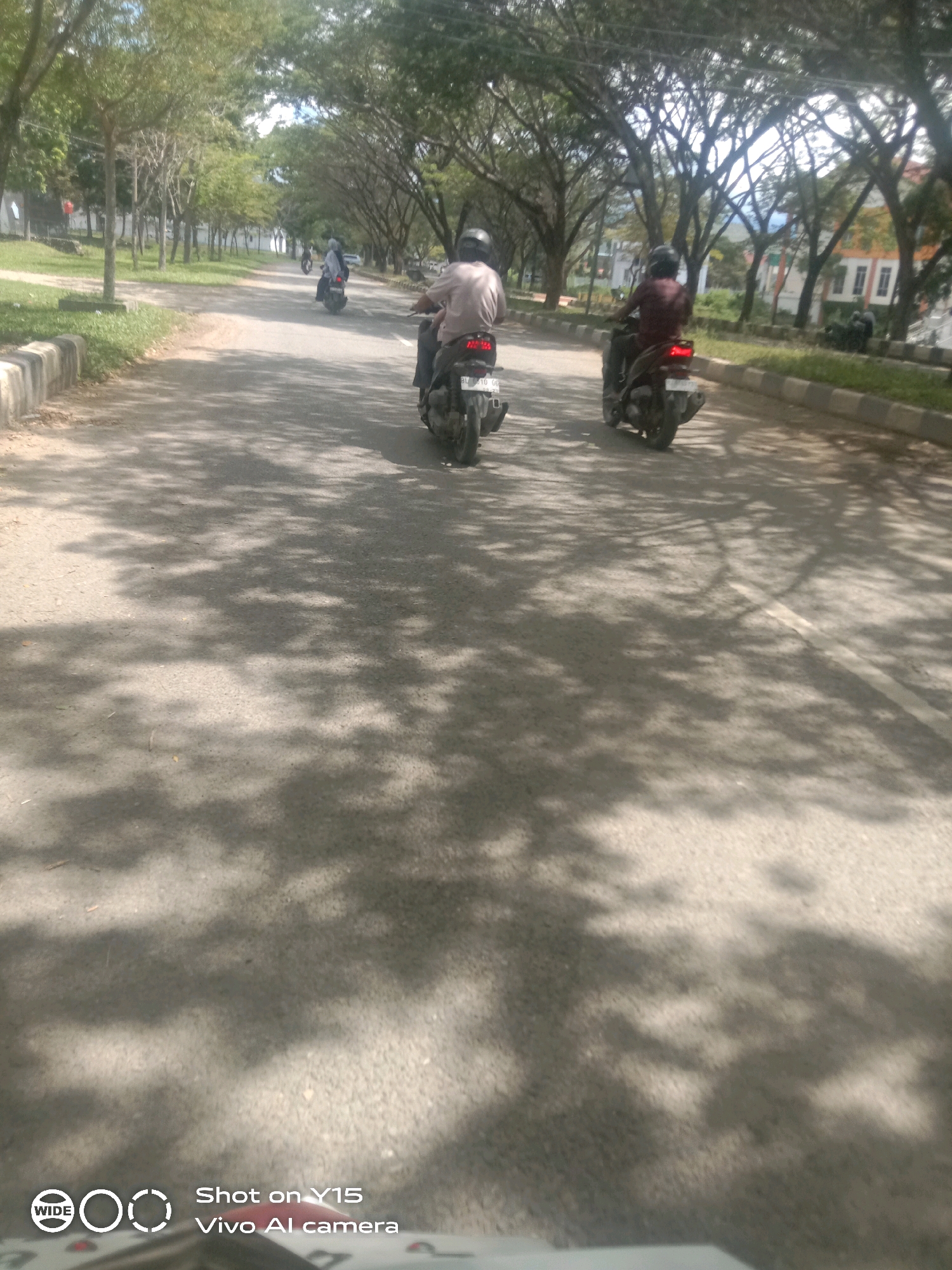 The image depicts a street scene in Meureudu, Indonesia, under a canopy of trees casting dappled shadows on the asphalt. Two individuals are riding motorcycles in the same direction on the road. The motorcycle in the foreground is ridden by a person wearing a light-colored shirt and a helmet. Behind them, another motorcycle is being ridden by a person wearing a darker shirt and a helmet. The road is lined with a curb and grass on the left side, and trees along the right. Buildings are visible in the background on the right side of the street. The overall impression is a typical sunny day in a town or city with tree-lined avenues.