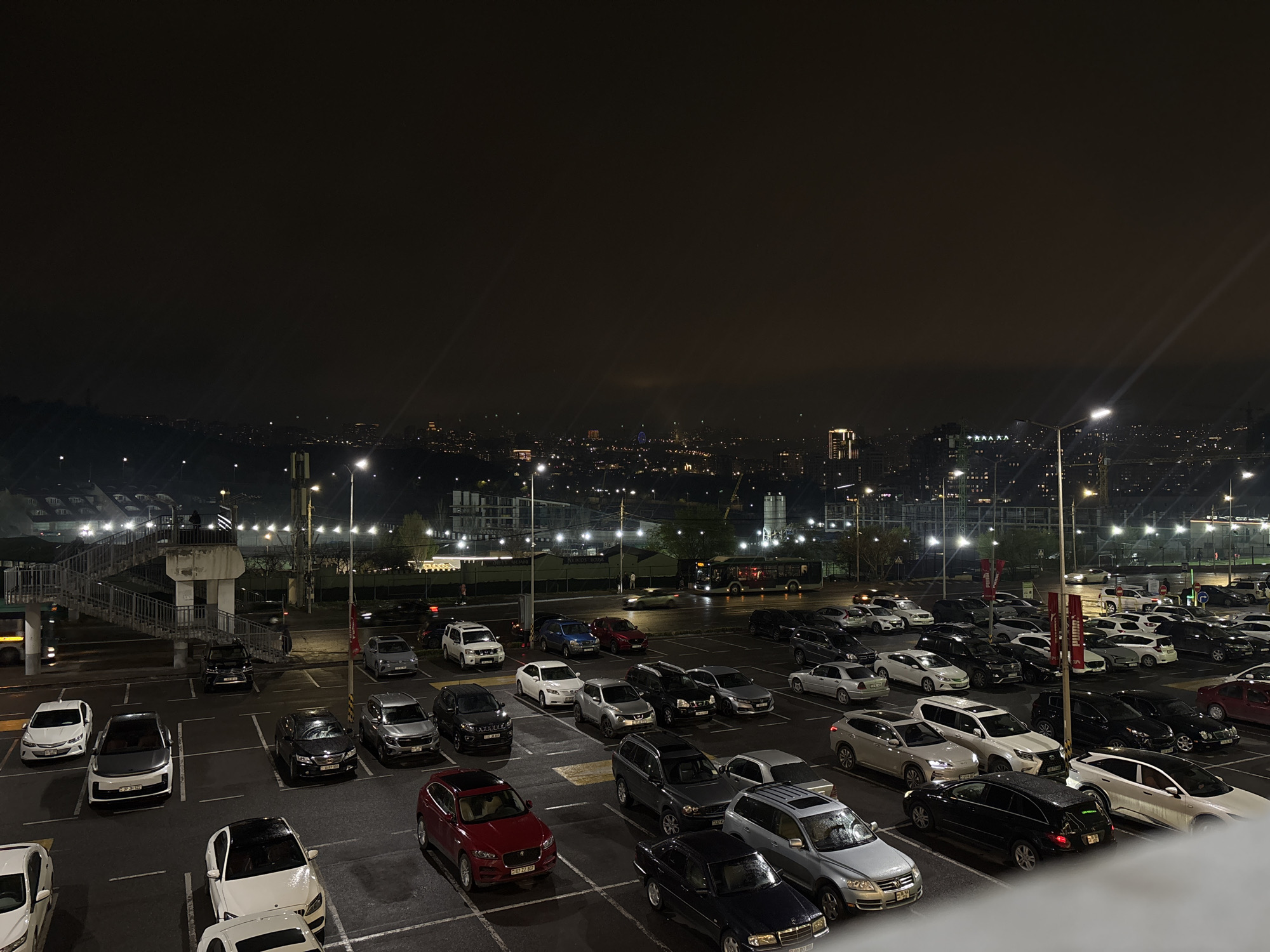 The image depicts an extensive parking lot filled with numerous vehicles at night, under the ambient glow of streetlights. In the background, the Yerevan city skyline is faintly visible, illuminated by distant city lights. A pedestrian overpass bridges the parking area. The parking lot surface appears wet, likely from recent rain. There are no clearly visible people in the immediate foreground. The environment is urban and likely adjacent to a large venue.