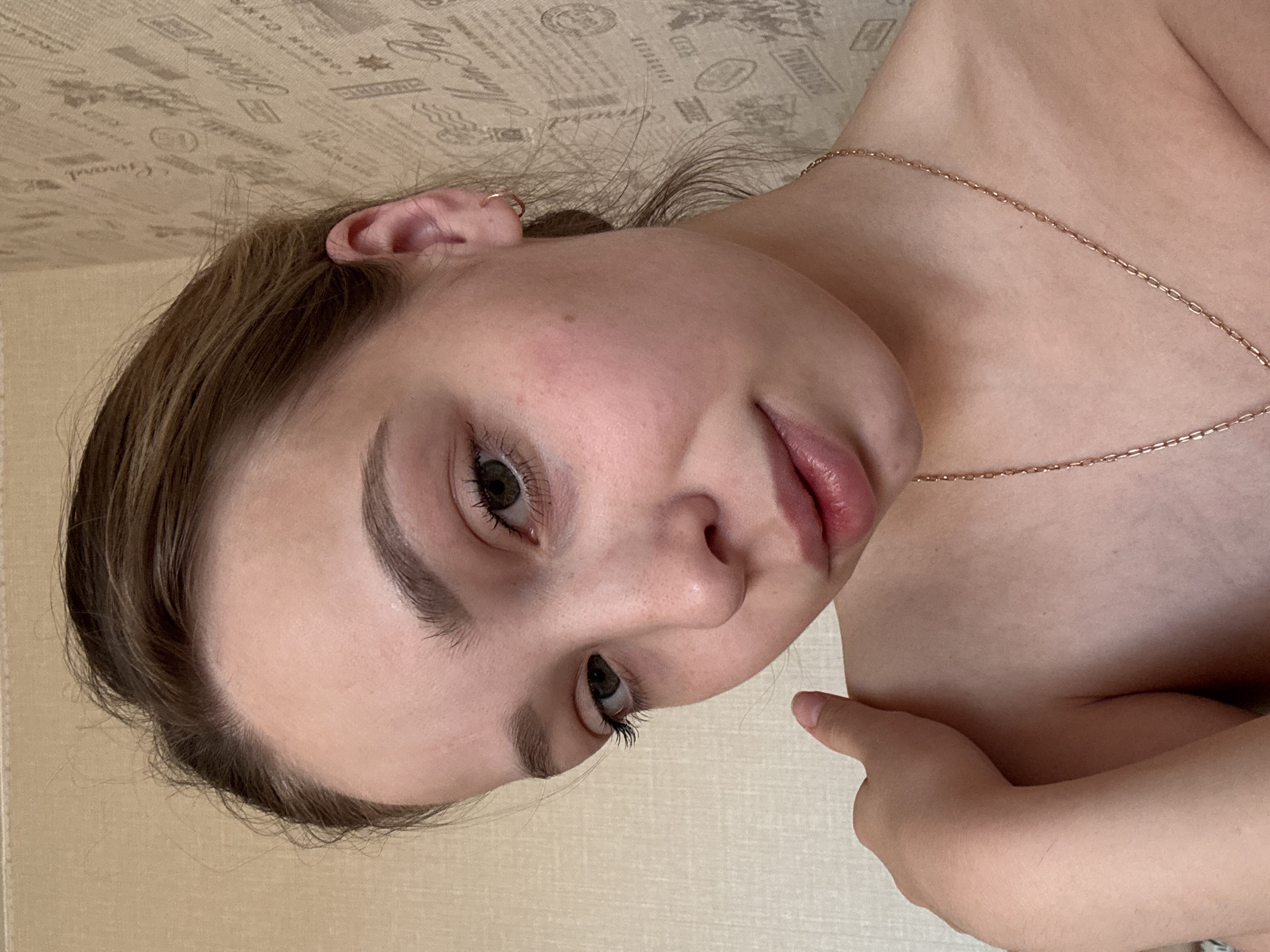 A close-up, angled portrait features a young woman with light to medium brown hair, which appears to be styled casually. Her head is tilted, and she gazes directly or slightly off-camera with dark eyes and defined eyebrows. Her lips are full and slightly parted, showing a neutral to mildly pensive expression. She has fair skin, with a small mole visible on her forehead.
The woman is adorned with a thin gold chain necklace and a small gold hoop earring in her visible left ear. Her bare shoulders and collarbone are prominent, suggesting she might be wearing a strapless top or is partially unclothed. Her right arm is bent, with fingers resting near her left shoulder or chest.
The background consists of a light beige or off-white wallpaper patterned with what appear to be various printed texts, stamps, and small illustrations, reminiscent of old documents or newspaper clippings. Visible English words on the wallpaper include "Crushed," "Shipping," "Coffee," "Original," and "TEA." The lighting is soft, suggesting an indoor setting, possibly a bedroom or living space. The image is taken in Chaikovsky, Russia.