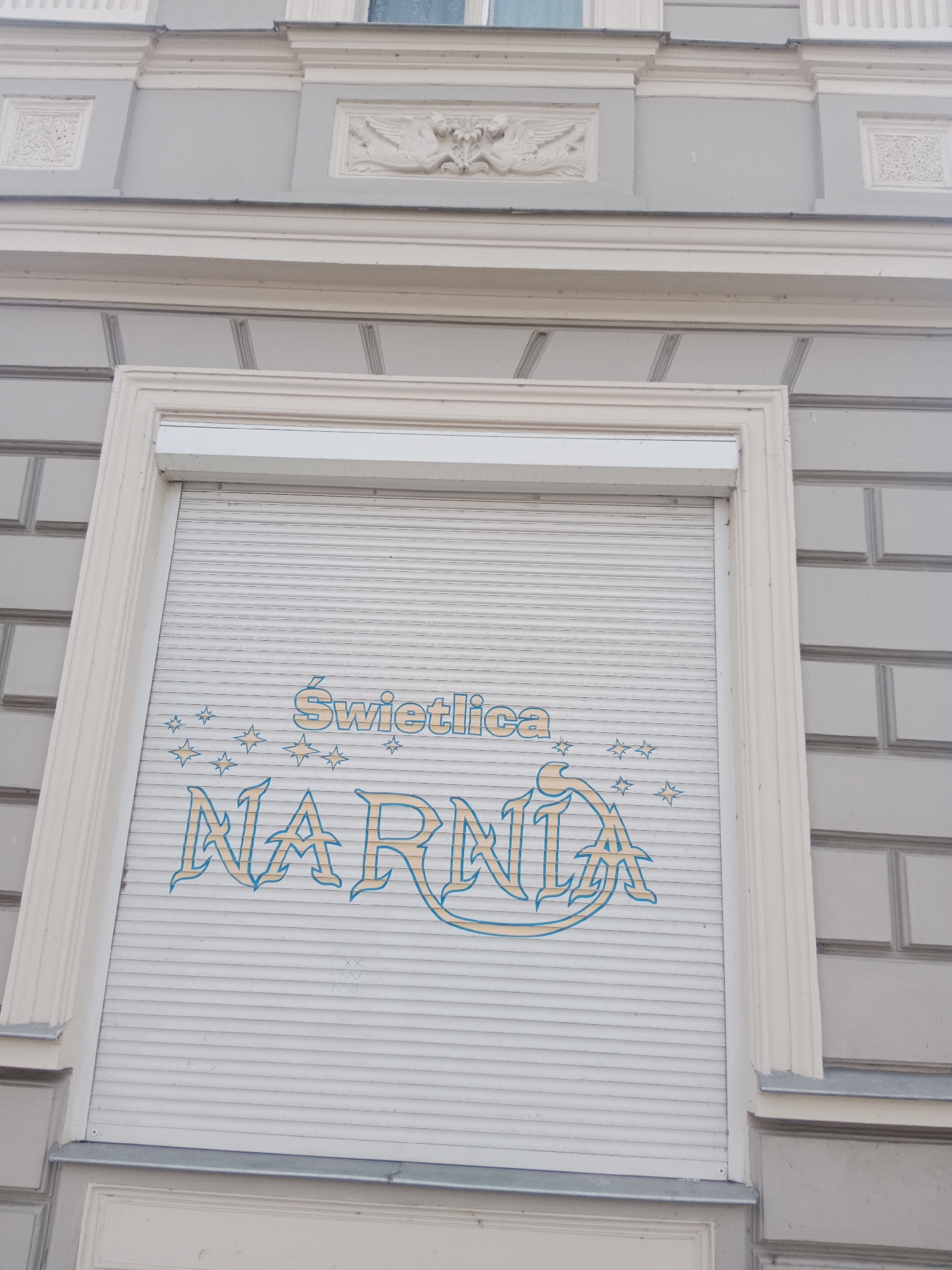 The image shows a close-up of a building facade in Bydgoszcz, Poland, featuring a closed white roller shutter with blue and yellow text and star graphics. The text on the shutter reads "ŚWIETLICA NARNIA". The architectural style of the building suggests it is an older structure, with decorative elements above the shuttered window. The surrounding wall is made of grey brickwork. The lighting appears to be natural daylight, and the image is taken from a slightly upward angle.