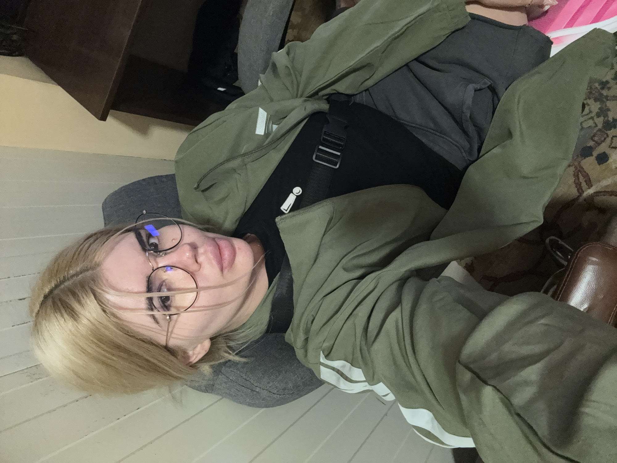 A young woman with blonde hair and glasses is sitting in a chair, wearing a green jacket over a black shirt and gray pants. She is looking at the camera with a neutral expression. The background shows a wall with vertical wooden panels and a desk. The image is a selfie taken from a high angle. The overall setting appears to be an indoor space, possibly a home or office, in Rakovski, Bulgaria.