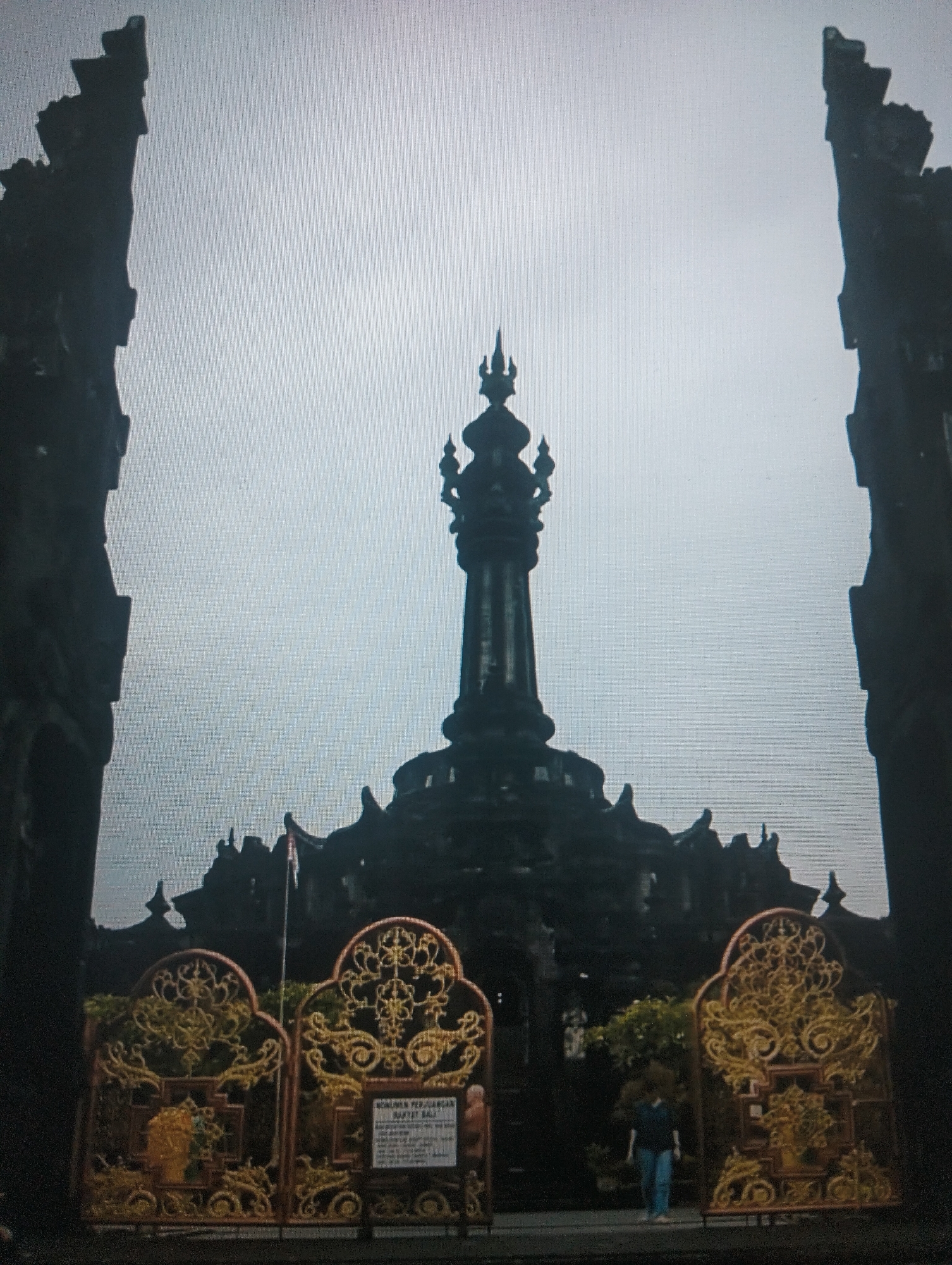 An imposing dark stone monument, topped with spires, dominates the center of the frame against an overcast sky. The monument is flanked by two tall, dark, silhouetted structures. In the foreground, ornate golden gates with intricate scrollwork are visible. A white sign is attached to the right gate. Behind the gates, lush green foliage can be seen. A person, wearing a dark top and blue jeans, stands near the right gate, facing away from the camera. The location is the Monument Perjuangan Rakyat Bali in the city of Medan, Indonesia.