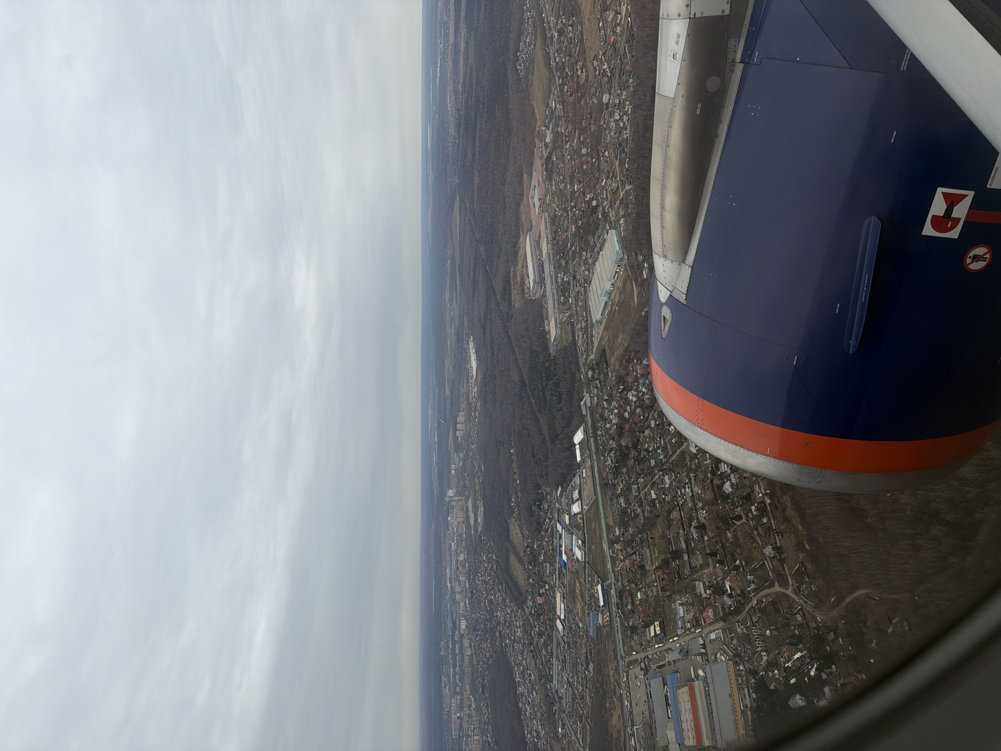 The image depicts a view from an airplane window, showing the wing and engine of the aircraft. Below, the city of Zaprudnya, Russia, is visible. The landscape is predominantly urban, with buildings and infrastructure spread across the area. A large expanse of forest is also visible. The sky is overcast and cloudy.