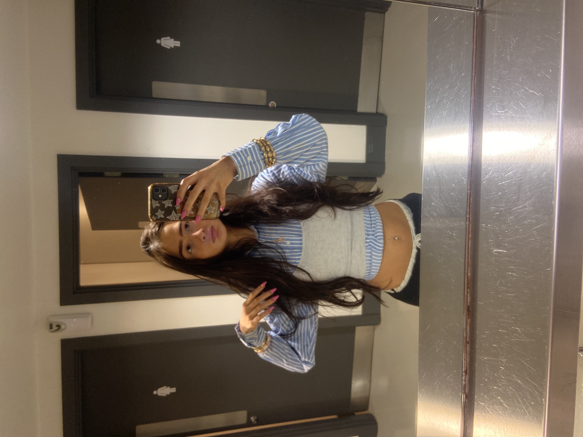 A woman is taking a selfie in a bathroom with stainless steel stall dividers. Behind her are two black doors with white restroom icons. The background wall is a light neutral color. The location is not discernible from the image.