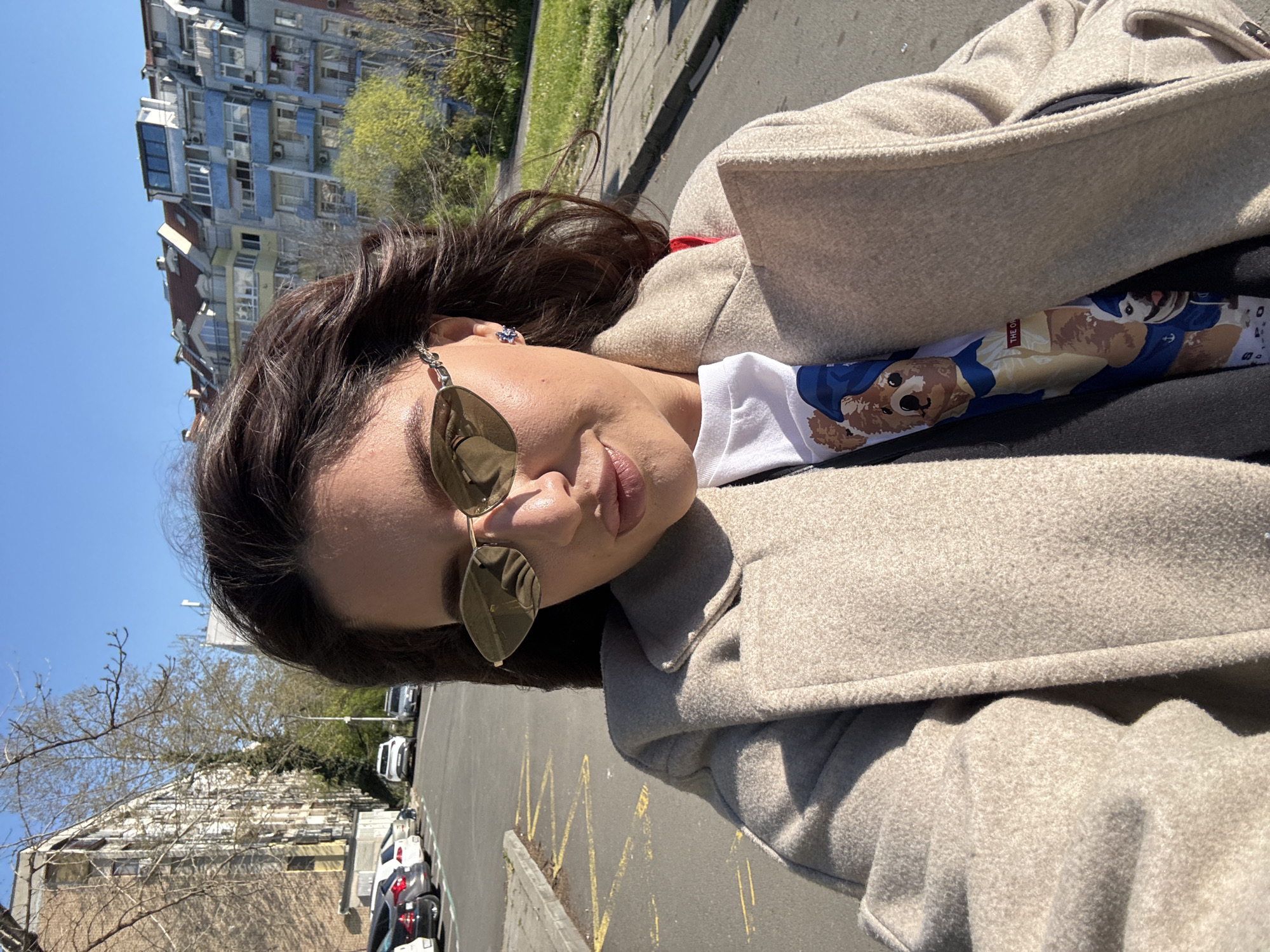 A woman wearing sunglasses and a beige coat takes a selfie in Burgas, Bulgaria. She has dark hair and is wearing a white shirt with a bear graphic. The background shows a city street with parked cars and apartment buildings. The sky is blue and sunny.
