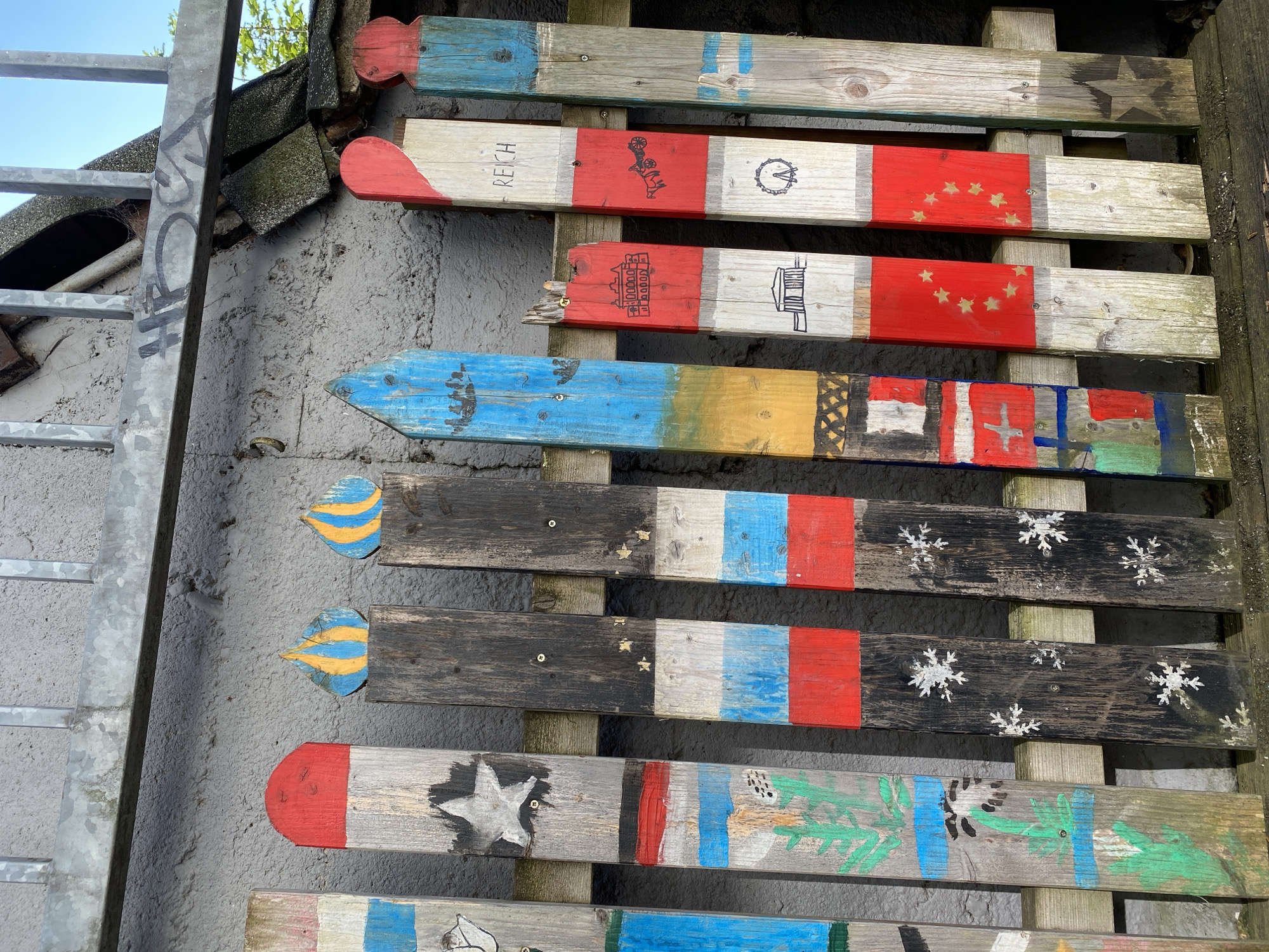 A series of wooden fence posts, painted with various colors and designs, stand in a row. The posts are of different heights and shapes, with some having pointed tops and others rounded. The paintings include flags, stars, snowflakes, and what appear to be depictions of buildings and a Ferris wheel. The posts are attached to a wooden railing and are set against a textured, grey wall. The scene appears to be outdoors, with natural light illuminating the objects. Some of the posts have visible screws or nails. A metal railing runs along the top of the fence. The image is taken from a slightly low angle, looking up at the fence. Based on external knowledge, this fence is located in Altenkirchen, Germany.
