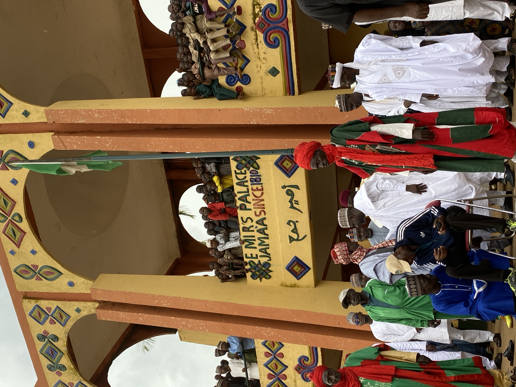 The image depicts a gathering of people in Kafanchan, Nigeria. Some individuals are observing from a structure with the inscription "EMIRS PALACE JAMAA SINCE 1810". A Nigerian flag is visible on a pole. Several men are standing or sitting, some wearing traditional garments with red turbans and green and red robes. Others are in casual attire, including an Adidas tracksuit. The structure is decorated with colorful patterns.