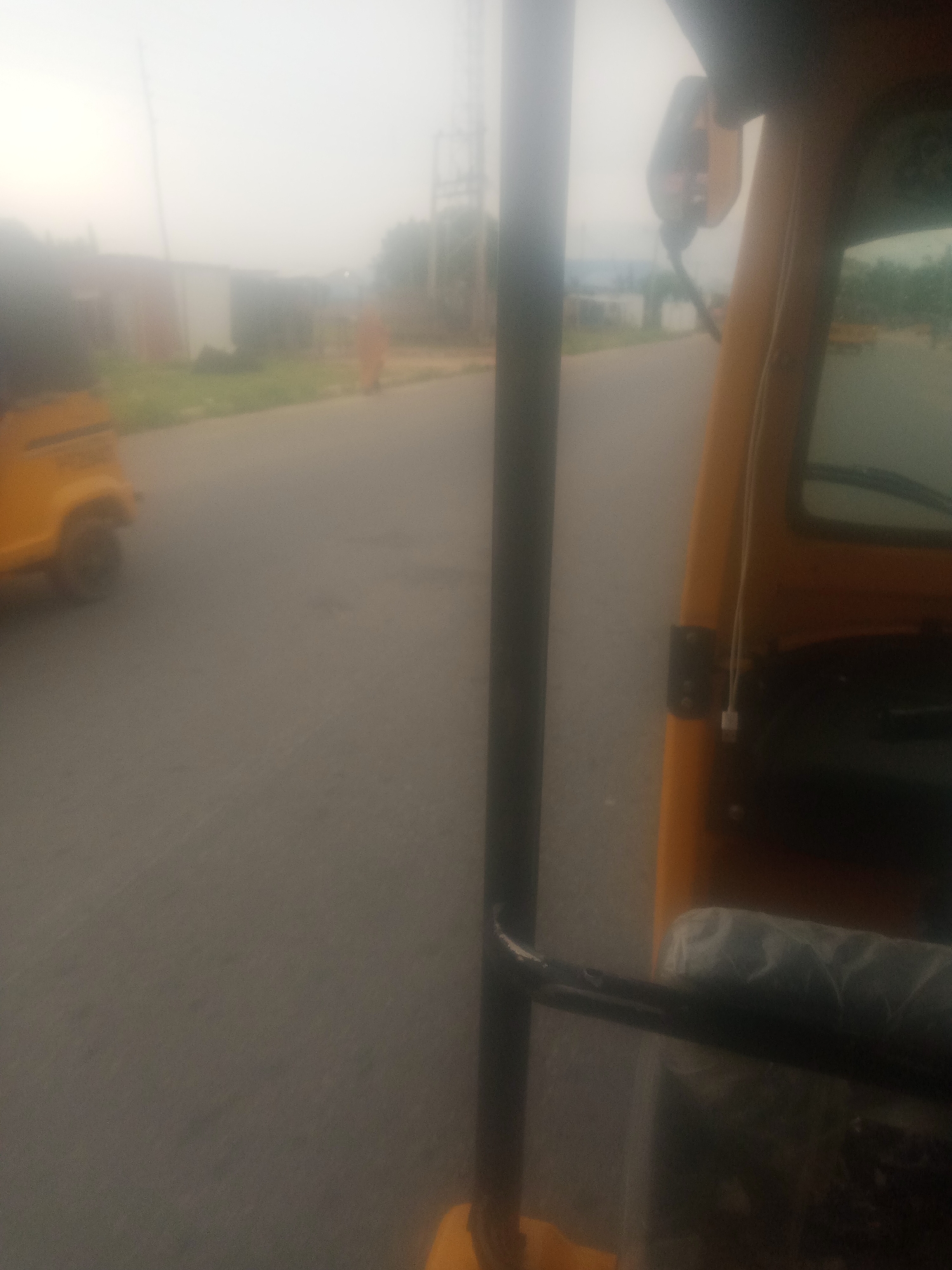 The image depicts a hazy, overcast day in Jalingo, Nigeria, likely on a street or road. In the foreground, a portion of a yellow auto-rickshaw, also known as a "Keke Napep", is visible on the right side of the frame. A dark vertical pole runs through the center of the image, obscuring a clear view of the scene beyond. To the left, another yellow auto-rickshaw is partially visible, appearing to be in motion. In the background, there are out-of-focus buildings and vegetation, and a person wearing an orange garment is walking on the side of the road. The overall atmosphere is muted due to the hazy conditions.