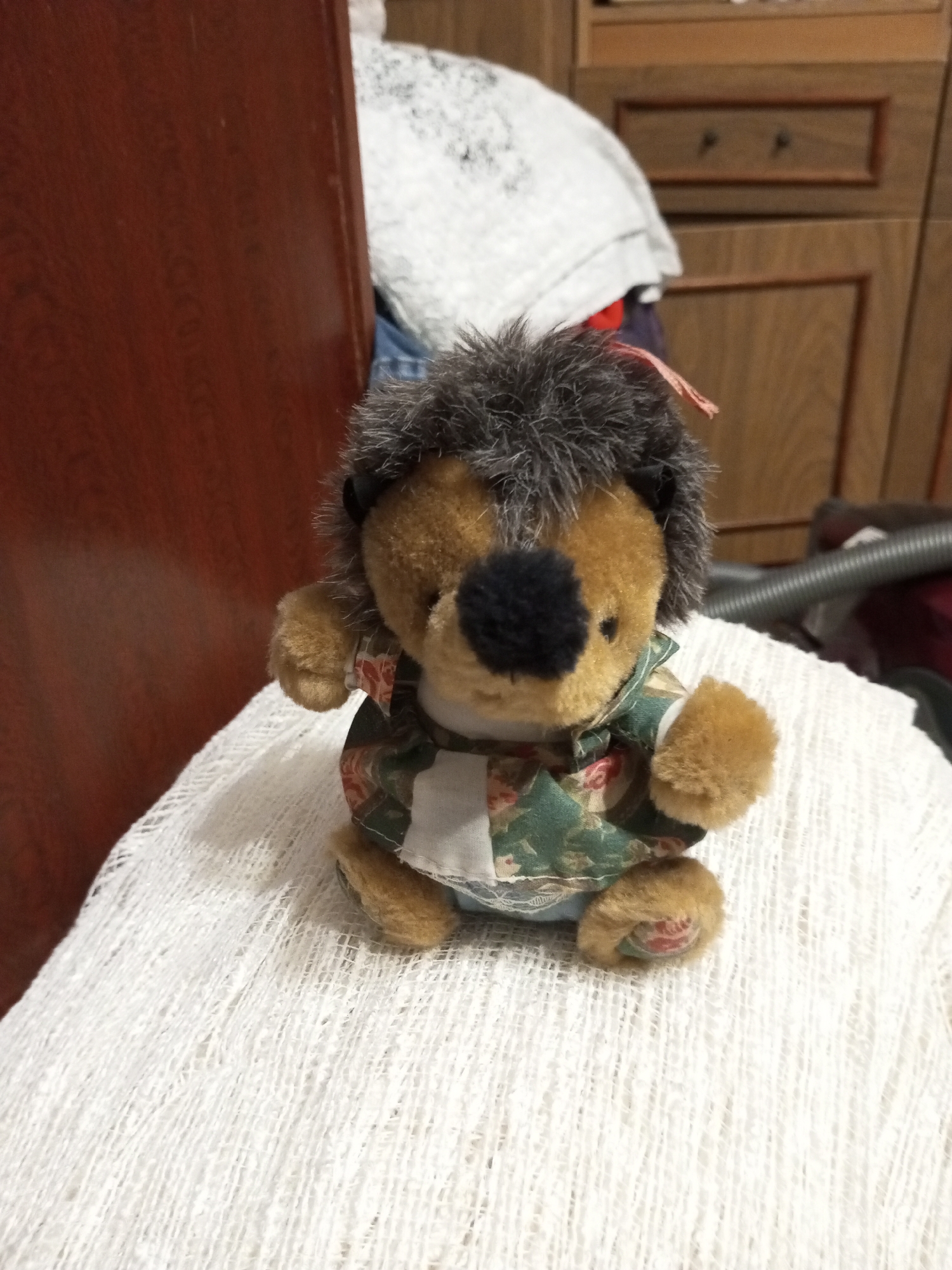 A plush toy hedgehog sits on a textured white fabric. The hedgehog has brown fur, a black nose, and grey, spiky "quills" on its head. It is wearing a green and white patterned shirt. The background is out of focus, but appears to be an indoor setting with wooden furniture and a white cloth. The scene is lit by artificial light. The location is specified as Ароматновское сельское поселение, Ukraine, but this information is not directly derivable from the visual content of the image itself.