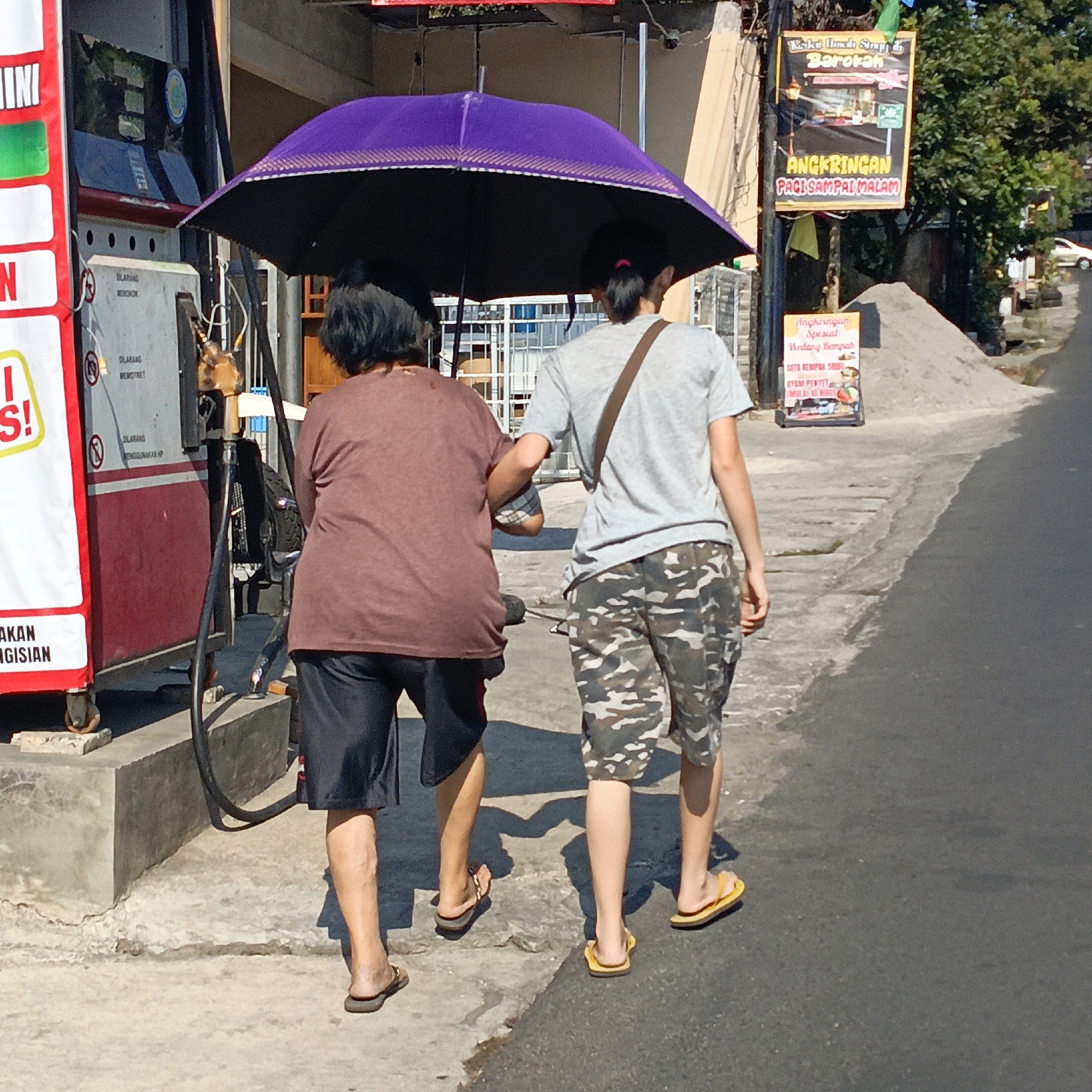 Two individuals, one appearing older and the other younger, are walking away from the camera under a large purple umbrella. The older individual, on the left, wears a brown t-shirt and dark shorts. The younger individual, on the right, wears a grey t-shirt with a brown strap across their chest and camouflage shorts. They are walking on a paved surface that transitions from a concrete sidewalk to an asphalt road.
Behind them is a red and white gas pump. The pump has signage with Indonesian text, including "DILARANG MEROKOK" (No Smoking) and "DILARANG MENGISI" (No Filling). Further in the background, a billboard-like sign displays "ANGKRINGAN PACI SAMPAI MALAM" (Angkringan until night), advertising a local food stall. To the right of the sign is a pile of sand and a tree. The overall environment suggests a street scene in a town or city. The presence of the gas station and the signs in Indonesian indicate the location is likely in Indonesia, possibly in the city of Salatiga, as suggested by the context of the request. The lighting suggests it is daytime, with shadows cast on the ground.