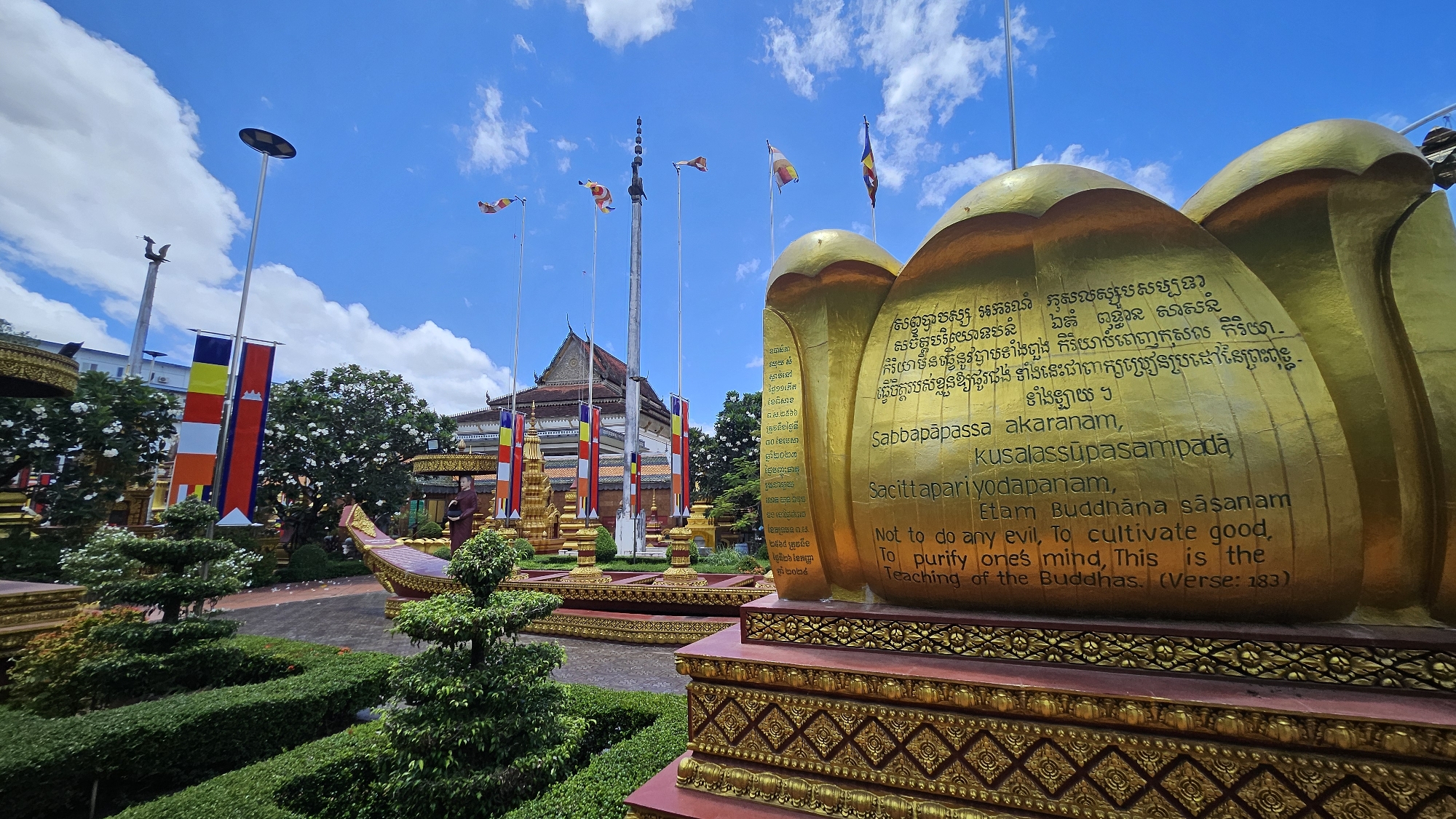 The image captures a serene Buddhist temple complex in Siem Reap, Cambodia, bathed in the bright light of a partly cloudy sky. In the foreground, a large, golden sculpture resembling a lotus flower is prominently displayed, inscribed with text in both Khmer script and English. The English text reads: "Sabbapapassa akaranam, Kusalassupasampadā Sacittapariyodapanam, Etam Buddhana sasanam Not to do any evil, To cultivate good, To purify one's mind, This is the Teaching of the Buddhas. (Verse: 183)".
Behind the golden sculpture, a row of colorful prayer flags, predominantly red, yellow, blue, and white, flutter from tall poles. The flags bear symbols and patterns indicative of Buddhist tradition. To the left, another set of flags, including the Cambodian flag, are also visible.
The scene is meticulously landscaped with neatly trimmed green hedges forming intricate garden designs. Several ornate, golden structures, possibly stupas or altars, are strategically placed within the garden. A traditional Cambodian boat-like structure, also adorned with golden embellishments, is situated in the middle ground. A statue of a robed Buddhist monk stands near this boat, adding to the spiritual ambiance of the complex.
In the background, a temple building with a distinctive tiered roof and ornate architectural details is visible, hinting at the religious significance of the location. The overall impression is one of tranquility, devotion, and cultural richness, characteristic of Buddhist sites in Cambodia.