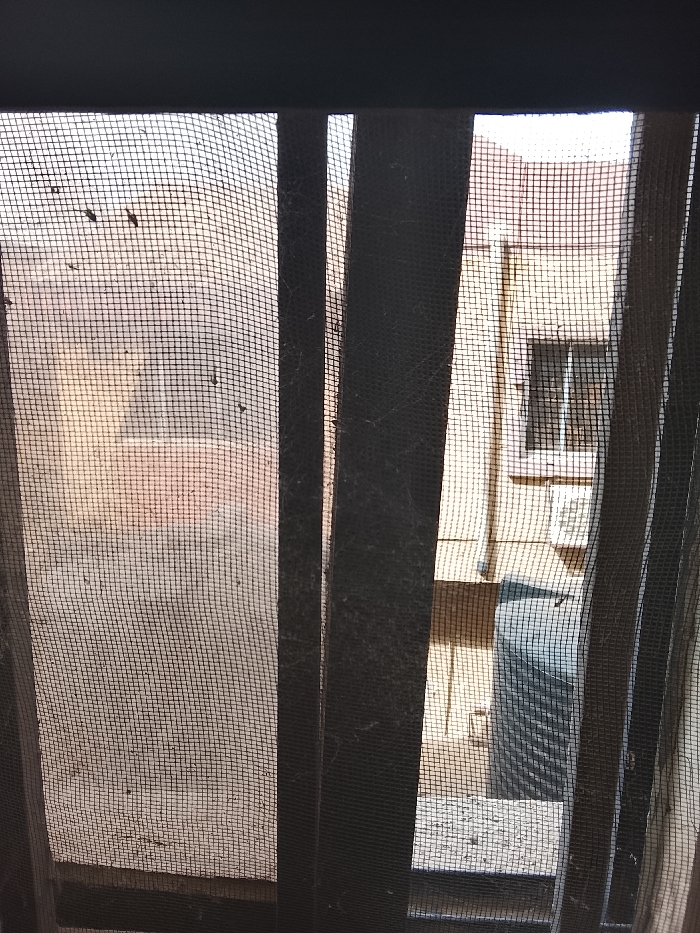 The image depicts an outdoor scene in Mgbidi, Nigeria, viewed through a heavily soiled and partially torn window screen. The screen occupies the entire foreground, characterized by a dark mesh fabric exhibiting numerous small debris particles, possibly dirt or insect remnants, along with visible cobwebs, particularly on the dark vertical bars of the window frame.

Through the screen, a light-colored building, likely residential, is visible in the midground. The building appears to have a textured surface, possibly painted concrete or stucco, with sections displaying a faded yellowish-brown hue, some with orange or pink undertones. A rectangular window with a distinct white frame is present on the building's facade, positioned slightly to the right of the central vertical screen bar. Below this window, a rectangular object, possibly an air conditioning unit or utility box, is affixed to the wall.

Further down and to the right, a dark blue or grey cylindrical object, resembling a barrel or drum, is partially visible on the ground level. A flat, light-colored surface, consistent with a concrete slab or paved ground, is visible beneath the building structures. The lighting suggests daylight conditions, casting subtle shadows and illuminating the scene. No individuals are visible.