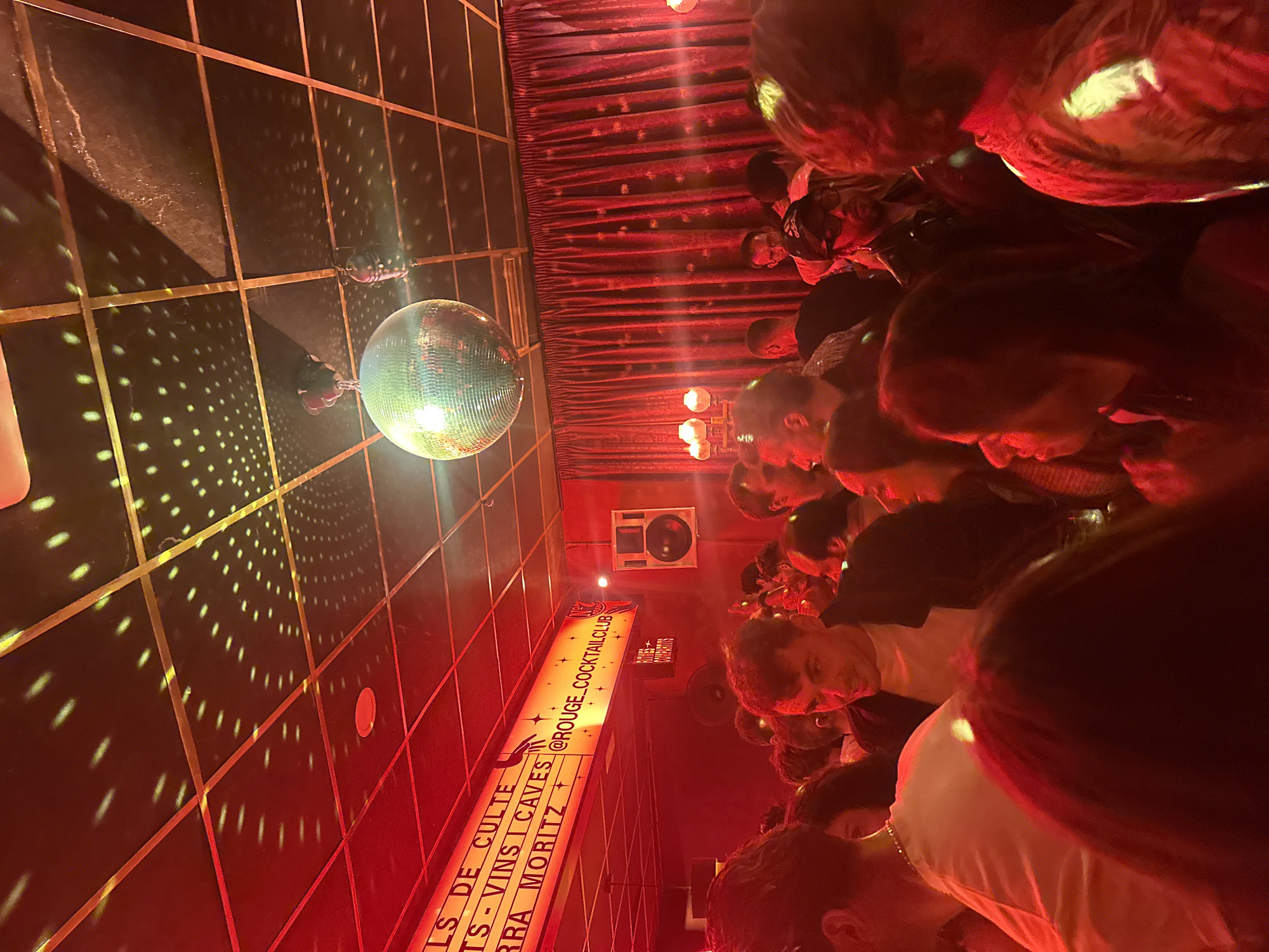 The image depicts a crowded interior space, likely a nightclub or bar, with a disco ball hanging from the ceiling. The room has a grid pattern on the walls and floors, illuminated by the disco ball's light. To the right, a large group of people is visible, densely packed together. Red lighting pervades the scene. On the wall, a sign reads "@ROUGE_COCKTAILCLUB" along with other text in French related to wines and cocktails. Based on the text "Moritz" on the sign, the location is likely in Barcelona, Spain, as Moritz is a popular beer brand from Barcelona. A speaker is mounted on the wall near the sign.