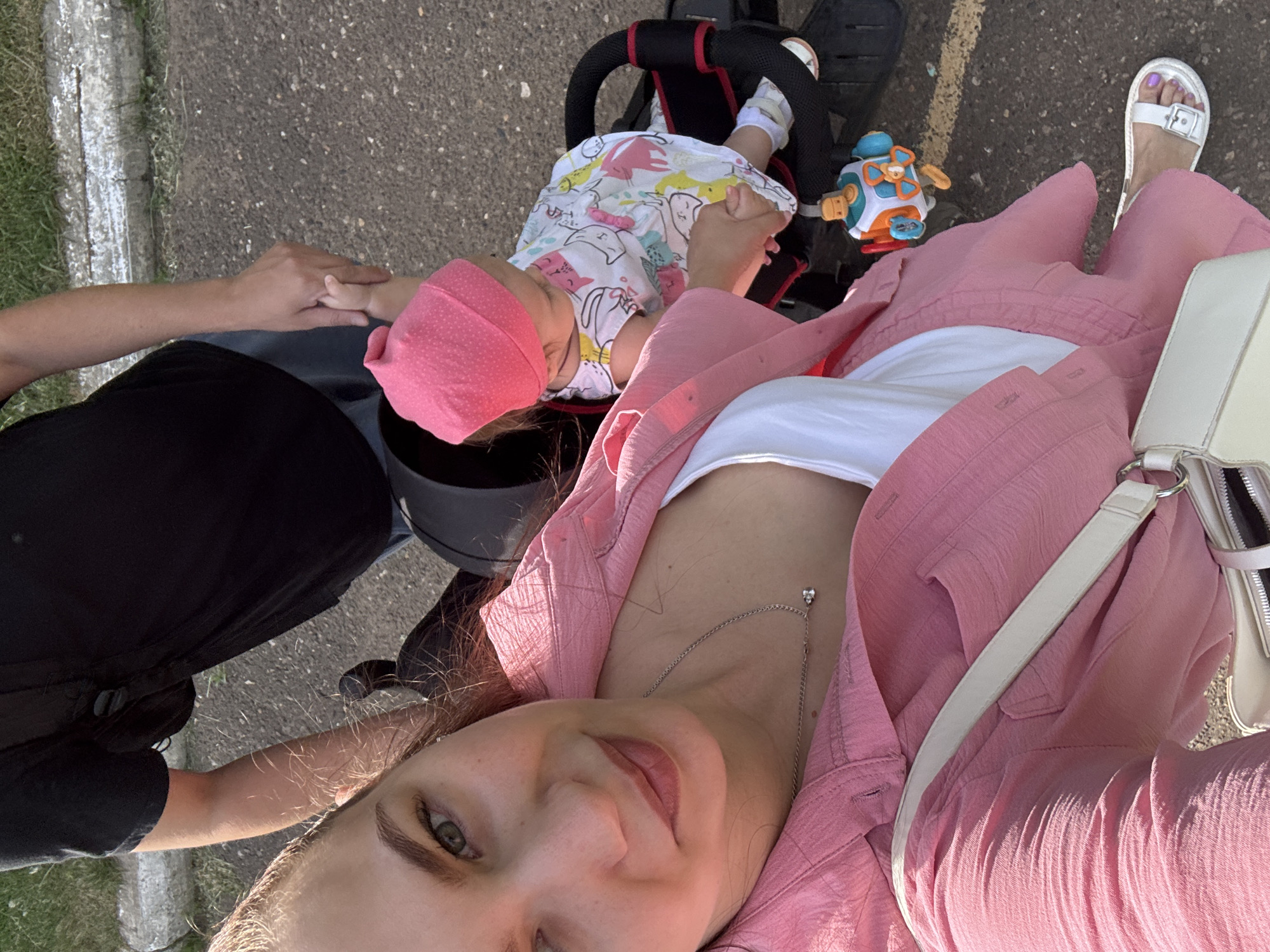 A selfie captures a woman in Chaikovsky, Russia, wearing a pink shirt and white top, holding a white purse. Her feet are clad in white sandals. Beside her, a child in a pink hat and printed shirt rests in a stroller. An adult male in a black shirt is seen holding the child's hand. A colorful toy is attached to the stroller. The ground appears to be paved, with greenery visible in the background, indicating an outdoor location in the city.