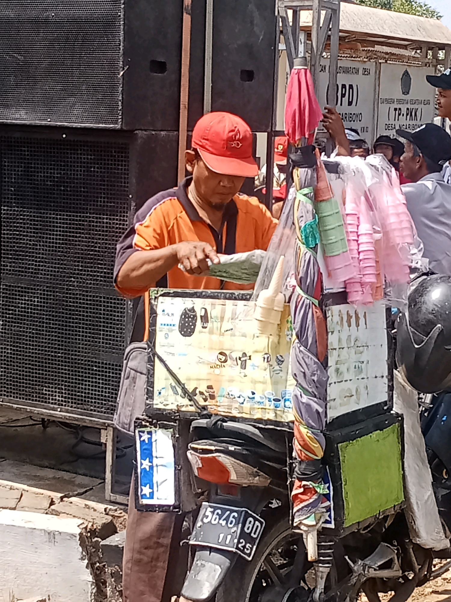 In the city of Ngariboyo, Indonesia, a street vendor is seen on a sunny day preparing to serve ice cream from his motorcycle. The vendor, wearing an orange shirt and a red cap, stands behind a meticulously organized cart attached to his motorcycle. The cart displays an array of ice cream cones and colorful cups, ready for customers. A prominent billboard in the background advertises the local government, "PEMERINTAH KABUPATEN KEDIRI (BPD) KECAMATAN NGARIBOYO", indicating the specific location. Several onlookers are gathered, observing the vendor's preparations. The scene captures a slice of daily life, showcasing the vibrant commerce and community spirit of Ngariboyo.