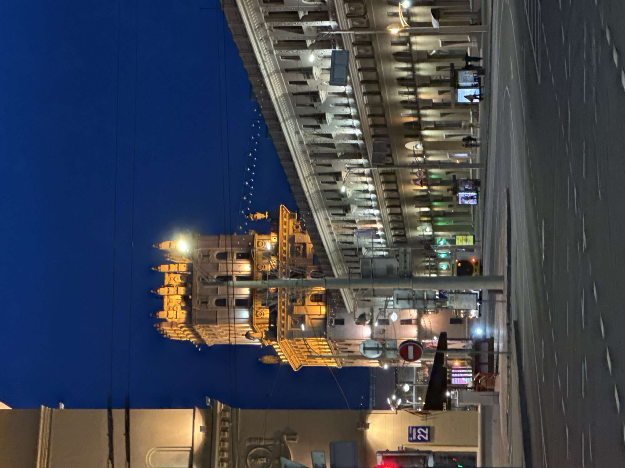 This nighttime image depicts a wide urban street scene in Minsk, Belarus, specifically at the intersection of Kirova Street, looking towards Independence Avenue. The sky is a dark, clear blue.
In the left-center background, a prominent multi-story tower, one of the "Gates of Minsk" buildings, is brightly illuminated with warm, golden light, accentuating its ornate Stalinist Empire-style architecture, including its decorative upper tiers and sculptural elements. To its right, a long, multi-story building, characteristic of Stalinist architecture along Independence Avenue, stretches across the mid-ground, also lit with warm uplighting highlighting its columns, balconies, and window recesses. A series of small, bright lights are strung horizontally in the dark sky above the street between these buildings.
The foreground features a wide, asphalt road with multiple lanes marked by dashed white lines. On the left sidewalk, a red traffic light is visible at the edge of the frame. A blue rectangular street sign with "22" and Cyrillic text, reading "УЛ КИРОВА" (Kirova Street), is mounted on a building. Adjacent to it, a small structure with a dark awning and illuminated interior is present. A red circular "No Entry" sign is prominently positioned on a pole in the middle of the scene. Further down the street on the right, a green traffic light is visible.
Several streetlights and utility poles are distributed throughout the scene. On the right side of the street, two modern bus shelters, each displaying illuminated advertisements, are visible. Indistinct figures of people are present near these shelters. Another individual stands near the small structure on the left. A vehicle with bright headlights is driving towards the viewer in the mid-ground, and a yellow bus is visible further down the road on the right. A small triangular speed limit sign indicating "40" is attached to the building on the right.