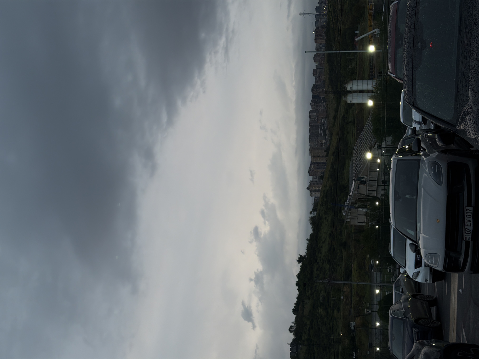 The image shows a line of parked cars in Yerevan, Armenia, under an overcast sky. The foreground is dominated by the vehicles, with a white car prominently displayed. Behind the cars, a green, sloping landscape leads up to a cityscape with buildings of varying heights. Streetlights are visible along the road and within the city. In the far distance, a power line tower stands silhouetted against the sky. The scene conveys a sense of urban life under a heavy, potentially rainy, sky.