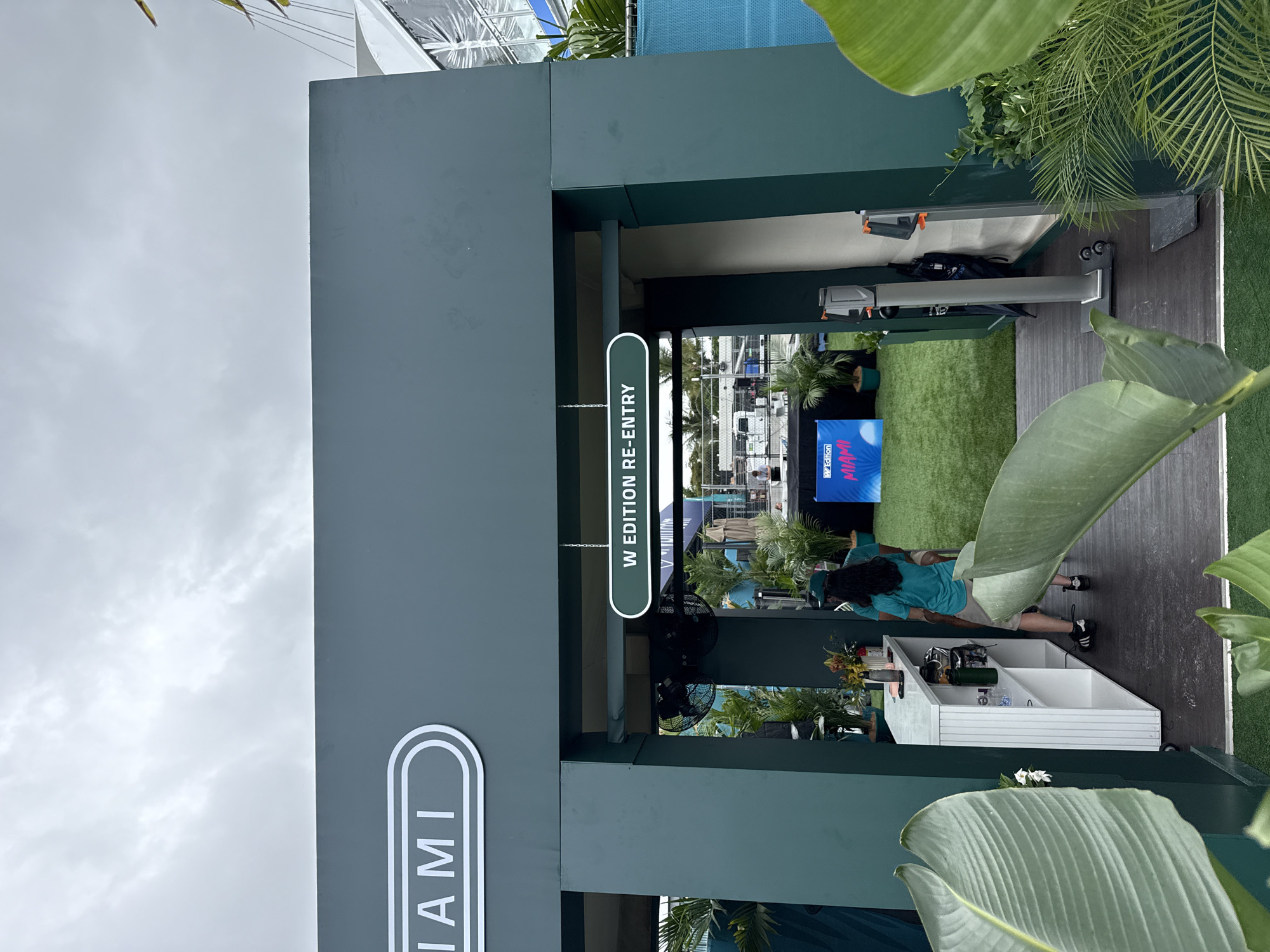 Here is a detailed description of the image:
The image shows an entrance at an event in Miami Gardens, United States. A large sign on the left reads "MIAMI" vertically. Above the entrance, another sign reads "W EDITION RE-ENTRY," indicating it is a re-entry point for the "W Edition" event. The structure surrounding the entrance is a dark green color.
Inside the entrance, there is artificial green turf. A television screen displays "MIAMI" with pink and blue accents. There is a person with dark hair wearing a blue shirt and shorts working at a white counter. The counter has several items on it.
The image is taken outdoors under an overcast sky. Lush tropical foliage is visible in the foreground and around the entrance, enhancing the Miami atmosphere. The overall scene is clean and well-maintained, suggesting a professional and upscale event.
