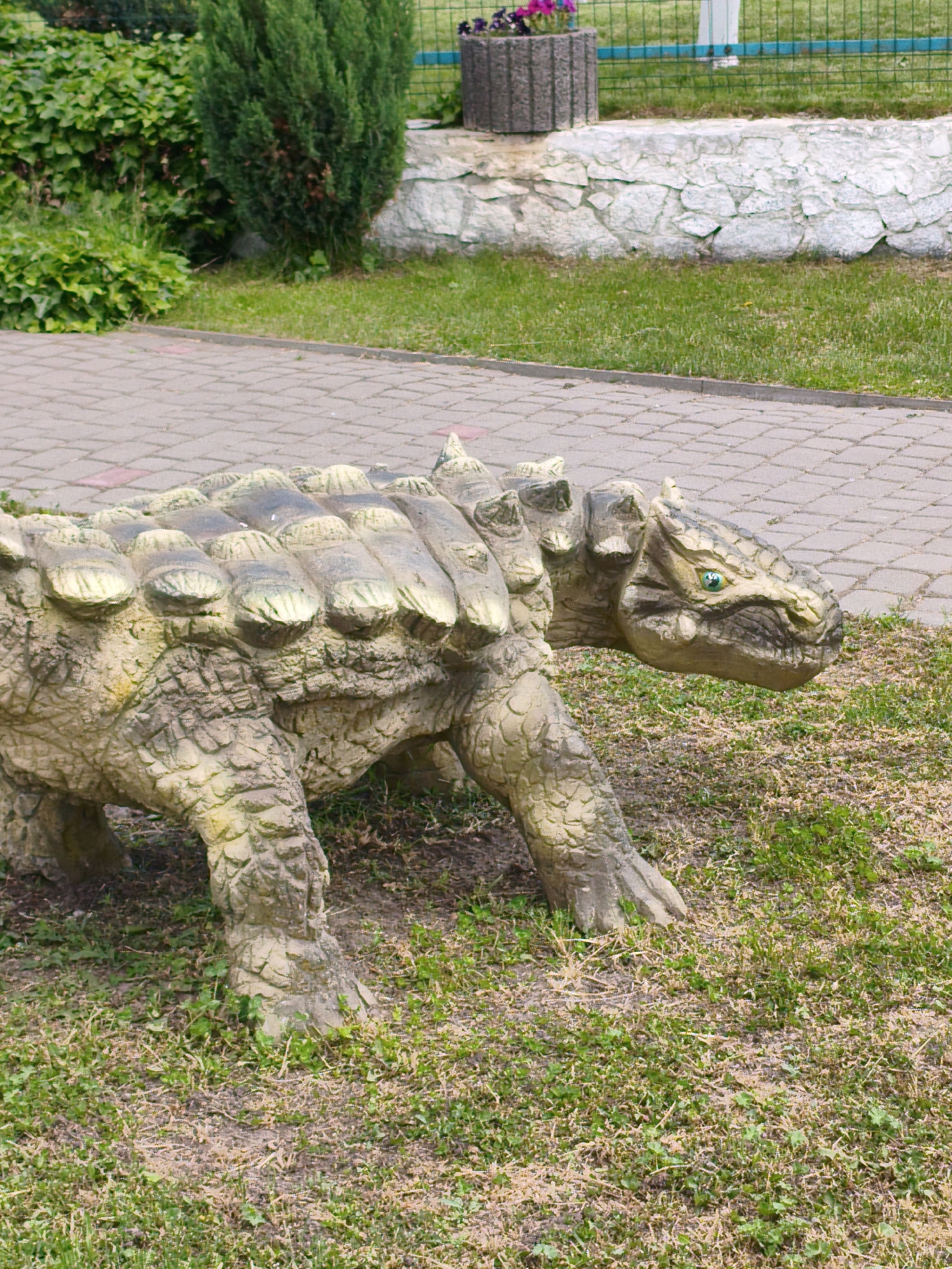 The image displays an outdoor scene featuring a large dinosaur statue in what appears to be a park or garden setting in Ustyvytsia, Ukraine.
The primary subject is a statue resembling an armored dinosaur, possibly an Ankylosaurus, positioned in the lower-left to mid-right of the frame. The statue is predominantly light beige or yellowish-brown with darker green and grey accents, suggesting age or weathered texture. It features a robust, scaly body with prominent, ridged armor plates or osteoderms covering its back and head. Its head is turned to the right, revealing striking green eyes. The dinosaur appears to be in a walking or standing pose, with its stout legs firmly planted on the ground.
The statue rests on a grassy area, which shows patches of dry grass and some bare soil, particularly around its feet. Immediately behind and to the left of the statue is a paved pathway constructed from interlocking bricks, featuring a repeating pattern of reddish-brown and grey squares.
In the background, further beyond the pathway, there is a vibrant green landscape. To the left, dense green bushes and a taller, dark green coniferous shrub or tree are visible. To the right, there's a low, white-painted stone wall with an irregular, textured surface. Above and behind this wall, a metal fence with blue horizontal bars and green mesh is partially visible. A grey concrete planter containing purple flowers sits atop or near the wall.
The scene is illuminated by bright daylight, suggesting a clear and possibly sunny day. There are no people visible, and no discernible text is present in the image. The overall impression is one of a well-maintained public space or attraction, likely designed for recreation or education.