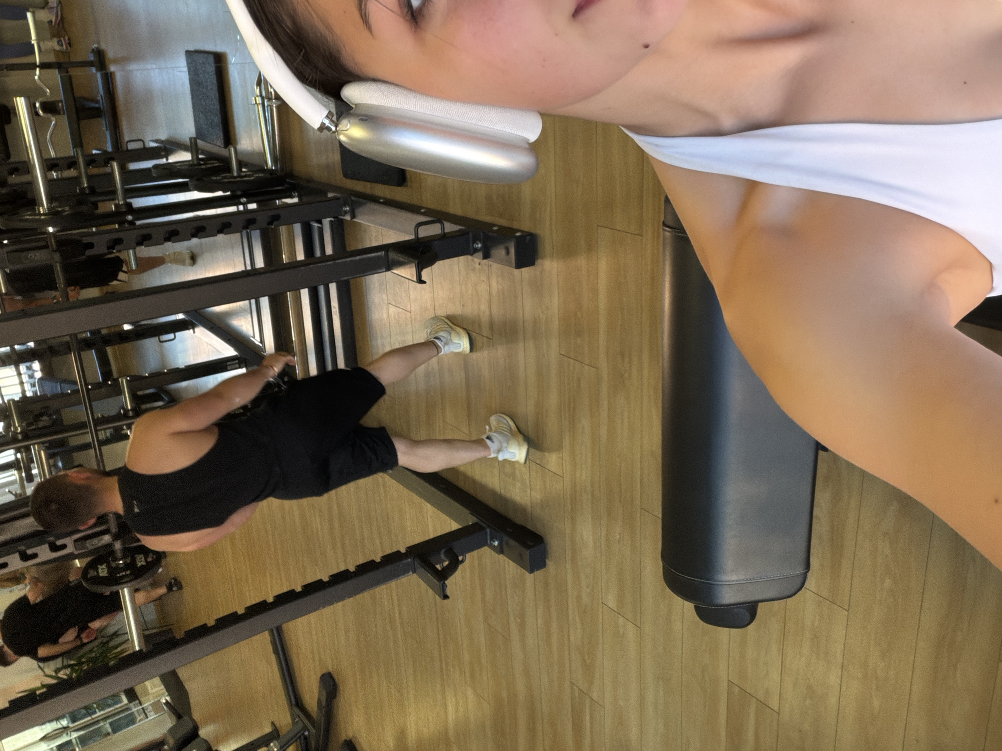 The image shows a person in a white top and headphones taking a selfie in a gym. In the background, another person is lifting weights. The gym has wooden floors and black workout equipment. The specific location in Gabrovo, Bulgaria, cannot be determined from the image.