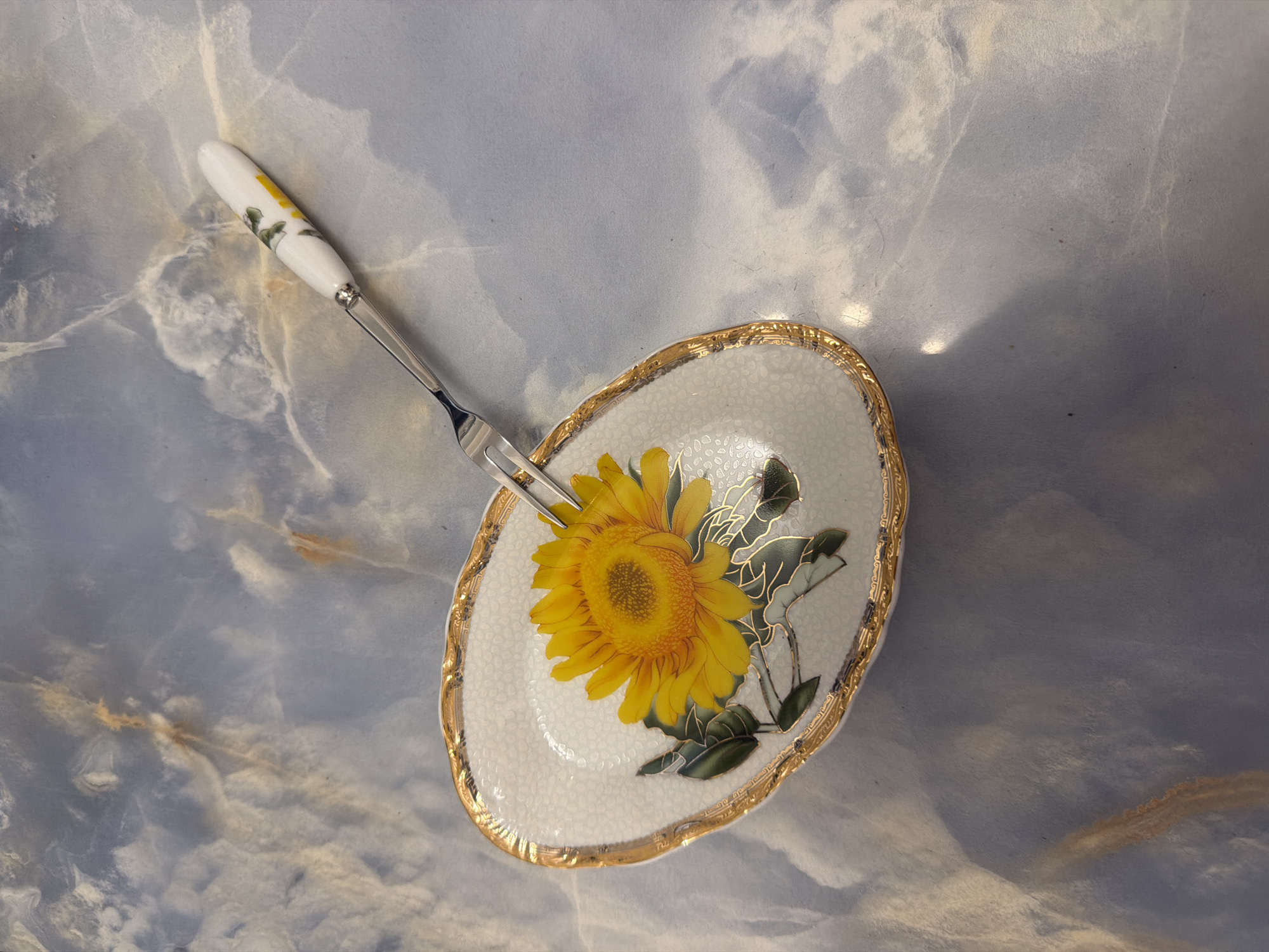 A single ornate pastry fork rests on a light blue, marbled surface. The fork has a white handle decorated with yellow flowers and a silver metal shaft leading to two tines. The tines of the fork are inserted into a small, oval-shaped ceramic dish. The dish has a white, textured interior and is adorned with a large yellow sunflower and green leaves. A gold-colored, decorative trim encircles the rim of the dish. The background is a soft, blue-gray surface with marble-like patterns of white and light brown.