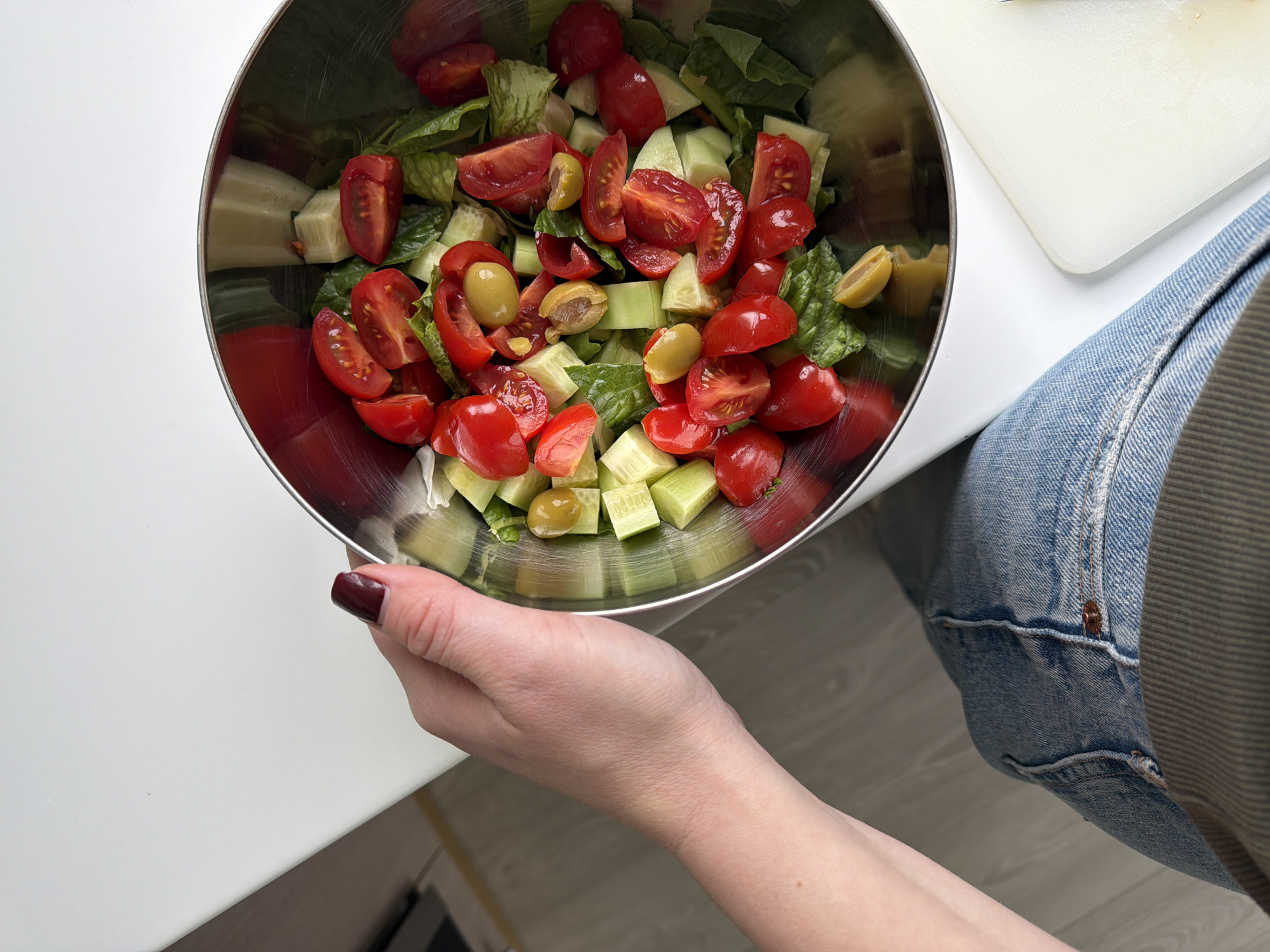 A photograph shows a partially filled stainless steel salad bowl containing mixed greens, halved tomatoes, cubed cucumbers, and sliced olives. The bowl is held in a person's hand with dark red nail polish. Next to the bowl is a white cutting board. The person is wearing blue jeans. The image appears to be taken indoors, with a white table surface visible in the lower left. The location is unspecified.