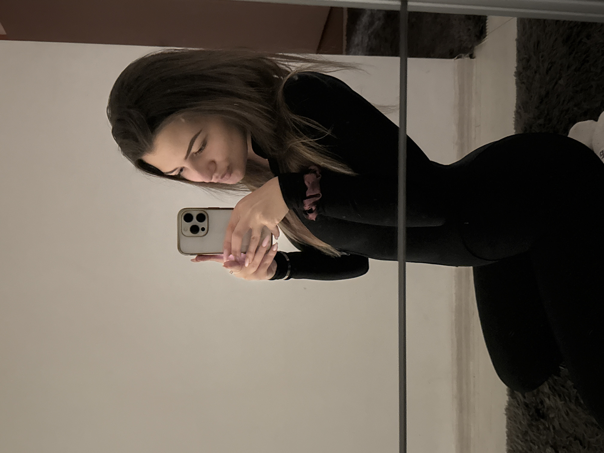 An indoor mirror selfie captures a young woman from the chest up, engaged in taking the photograph. The image is rotated 90 degrees counter-clockwise, showing her head on the left side of the frame, angled downwards, and her body extending to the right. She has long, dark brown hair and wears a form-fitting black long-sleeved top or jumpsuit. Her expression is focused or contemplative, as she looks down, seemingly at the screen of the light-colored smartphone she holds with both hands. The phone features a prominent multi-lens camera module. Her fingernails are painted a light pink or nude shade, and a ruffled pink/mauve scrunchie is visible on her left wrist or sleeve.
The background primarily consists of a plain, light-colored wall, contrasting with a dark, shaggy textured surface visible in the upper and lower right sections of the frame, likely a rug or carpet. A thin, dark vertical line, possibly a mirror edge or frame, runs through the image. The lighting is soft and even, characteristic of an indoor environment. The scene takes place in Sofia, Bulgaria.