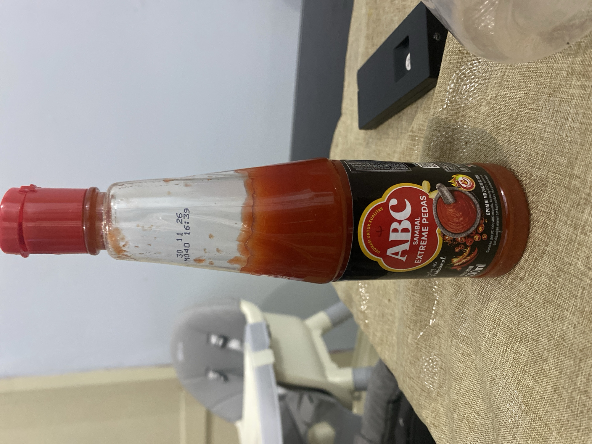 A tall, clear glass bottle of ABC Sambal Extreme Pedas hot sauce is centered in the frame, standing upright on a textured, tan-colored tablecloth. The bottle has a red cap and is filled with a vibrant red sauce, with some residue visible on the inside of the glass. The label is black with yellow and red accents, featuring the "ABC" logo and the text "SAMBAL EXTREME PEDAS." The date "30 11 26" and time "16:39" are printed on the glass near the neck of the bottle. To the left of the bottle, out of focus, is a gray baby high chair. To the right of the bottle, a black electronic device, possibly a remote control or a small case, rests on the tablecloth. The background is a plain, light-colored wall. The scene appears to be indoors, likely a home environment. The location of Dukuh, Indonesia, is not visually discernible from the image itself.