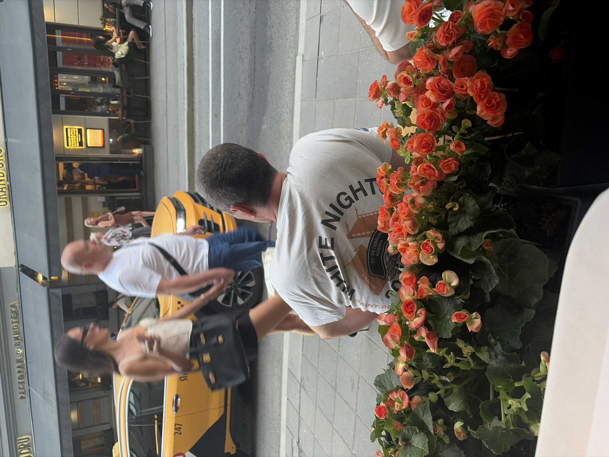 Here is a detailed description of the image:
**Scene Overview:**
The image captures a street scene next to a restaurant named "Grand Cru". A taxi is parked near the curb, and people are walking alongside it. The scene includes individuals in varying states of activity, from walking to standing.
**People:**
1. A man with short hair is wearing a white t-shirt with a graphic and lettering on the back. Part of the lettering reads "WHITE NIGHT" and "SAINT-PETERSBURG RUSSIA." He is standing near the taxi and facing a display of flowers.
2. Another man in a white shirt is walking with a black shoulder bag.
3. A woman wearing a light-colored top and a dark skirt or shorts is walking. She carries a black handbag.
4. Additional people can be seen seated at tables inside the restaurant.
**Objects:**
1. **Taxi:** A yellow taxi with "247" visible on its side.
2. **Flowers:** A large arrangement of bright orange flowers is situated near the sidewalk.
3. **Restaurant Sign:** The restaurant "Grand Cru" is visible with Russian text below.
4. **Furniture:** Tables and chairs inside the restaurant are visible through the windows.
**Activities:**
1. People are walking along the sidewalk.
2. People are seated inside the restaurant.
3. The taxi appears to be either parked or waiting.
**Environment:**
1. **Location:** The scene is on a city street adjacent to the "Grand Cru" restaurant.
2. **Weather:** The lighting suggests it is daytime.
**Overall Impression:**
The image captures a typical urban scene, potentially in St. Petersburg, Russia, given the lettering on the man's shirt. The presence of the taxi, people, and the restaurant all contribute to a dynamic snapshot of city life.