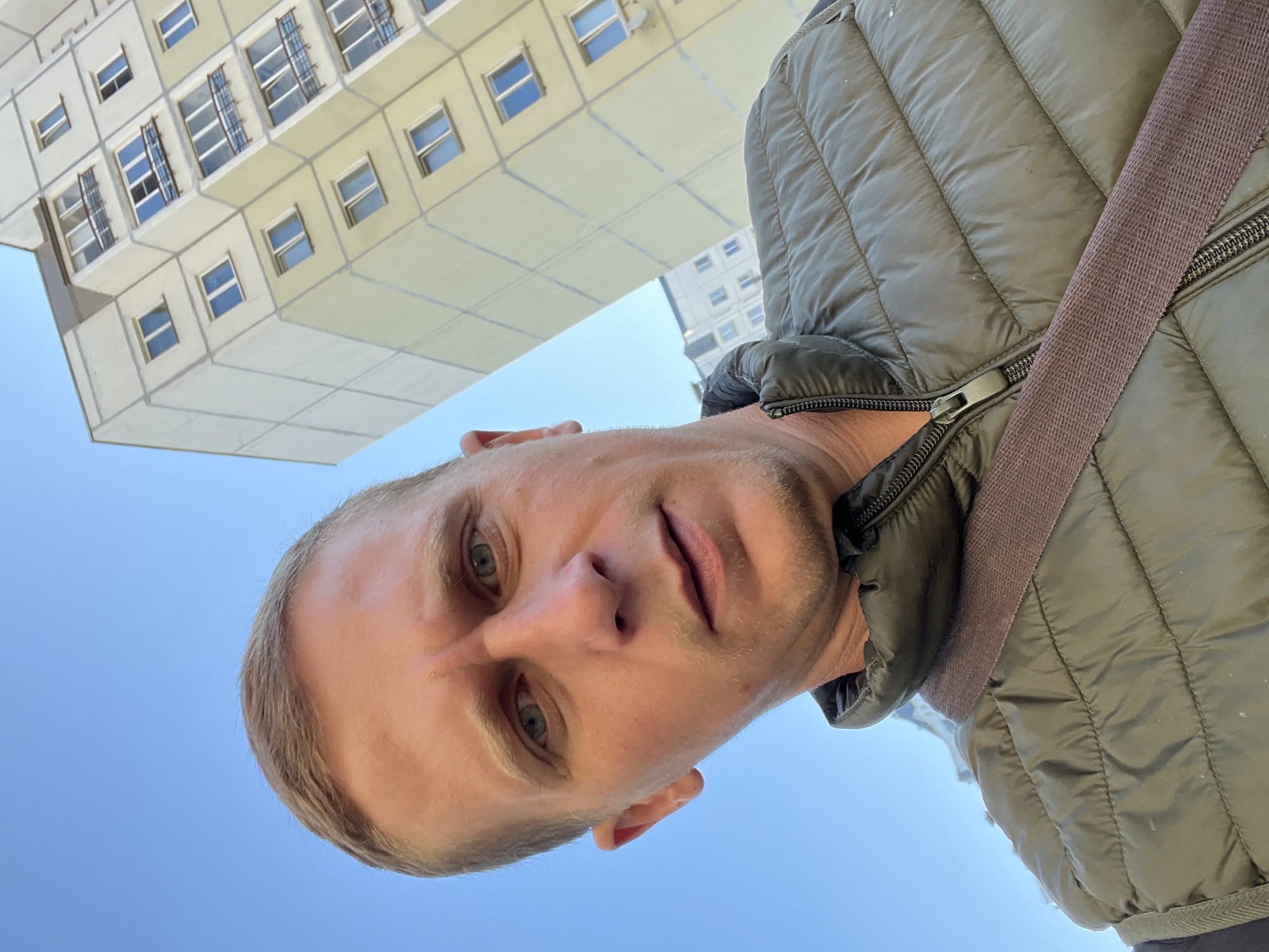 A selfie is taken outdoors in Minsk, Belarus, with a man in the foreground and a building in the background. The man, who appears to be in his 30s or 40s, is wearing a dark green puffer vest over a dark shirt and has short, light brown hair and blue eyes. He is looking directly at the camera. The building behind him is a multi-story apartment building with a yellow facade and numerous windows. The sky is clear and blue. The angle of the shot is from below, looking up at the man and the building.