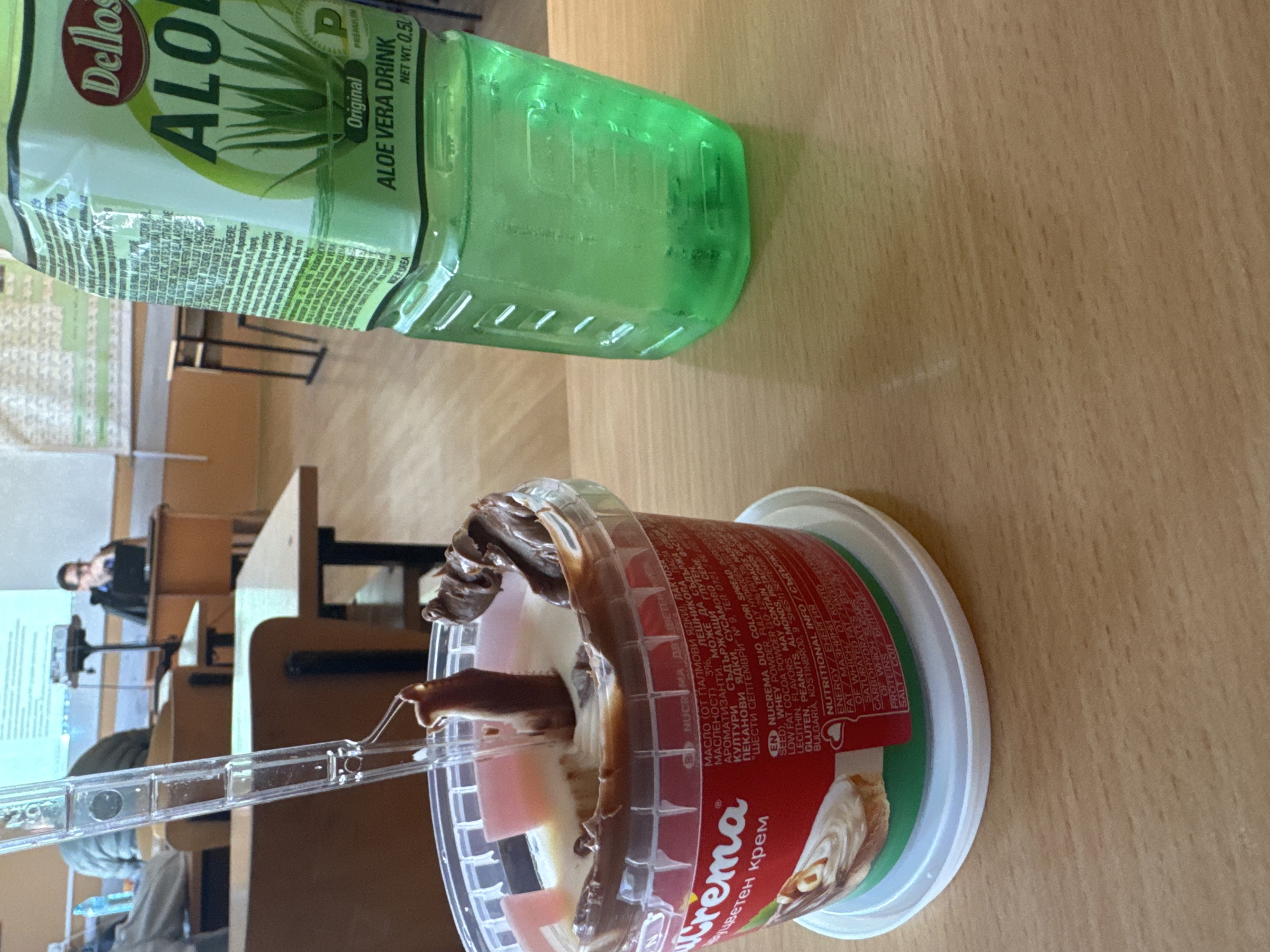The image shows a table with a bottle of Dellos Aloe Vera Drink and a container of Nucrema Duo Colori spread, along with a spoon. The Nucrema container label includes text in Bulgarian and references a location in Kostinbrod, Bulgaria at "Шести септември" № 9, implying this image could be from somewhere nearby such as Buhovo. There are people in the background of the image. The table is in a room with desks and chairs, suggesting a classroom or office setting.