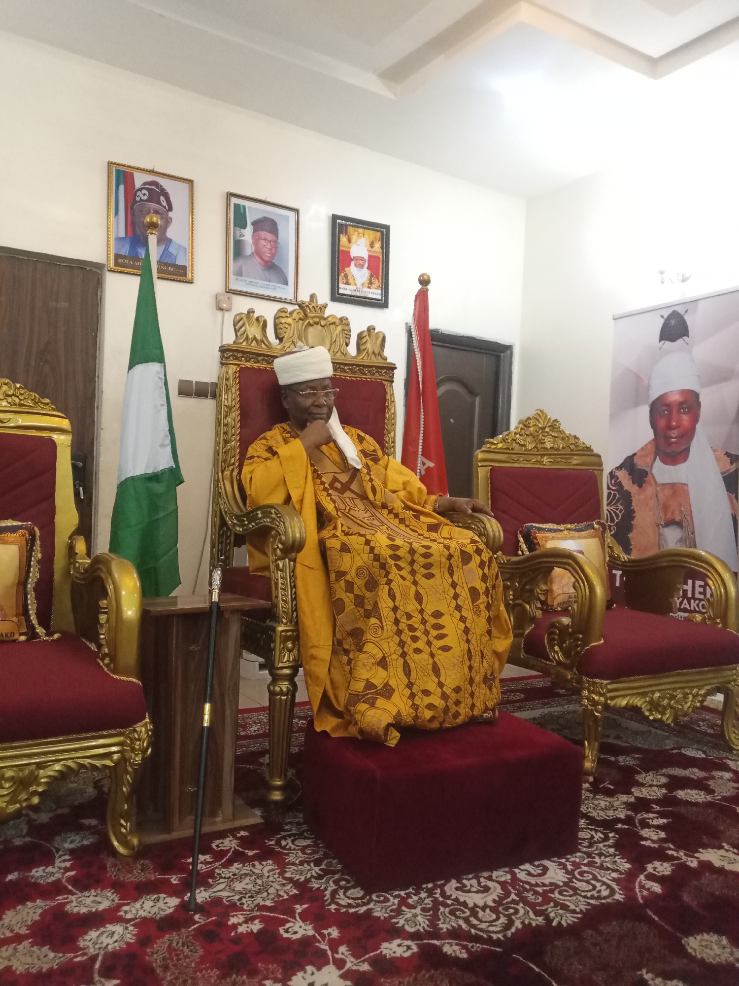 An elderly man sits on an ornate golden throne in a room setting, seemingly in a position of authority. The man is dressed in a mustard yellow robe with brown geometric patterns and a traditional white cap. He wears glasses and has a thoughtful expression.
To the left of the throne stands a Nigerian flag, and portraits hang on the wall above, possibly of historical or political figures. A walking stick leans against a small wooden table beside the throne. To the right, another throne is visible along with a large banner displaying a portrait of a man dressed in similar traditional attire.
The floor is covered with a detailed red and white patterned rug. The location appears to be indoors in a formal or ceremonial setting, likely in Unknown, Unknown.