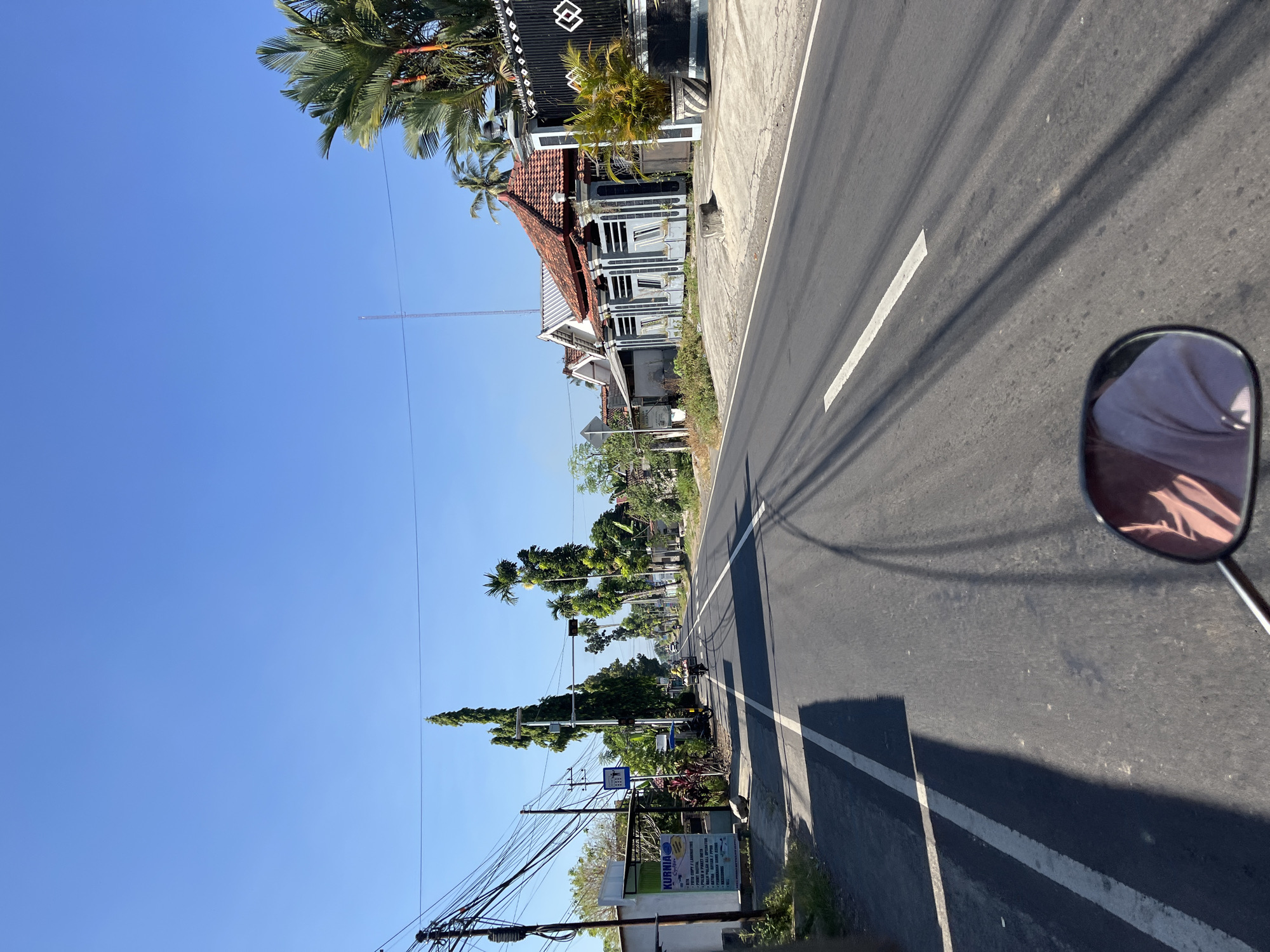 The image depicts a street scene in Senduro, Indonesia, on a clear, sunny day. The perspective is from a moving vehicle, likely a motorcycle, as indicated by the reflection of a person's torso in the lower right corner mirror. The road is paved and has a white line marking the center. On the left side of the road, there's a small building with a sign for "KURNIA". Overhead, a complex network of electrical wires crisscrosses the sky, supported by poles. Tall, slender trees line the street, interspersed with palm trees and residential houses with tiled roofs. Several of these houses feature decorative black fences and potted plants. A single person on a motorcycle is visible further down the road, moving away from the viewer. Traffic signals are also present along the street. The environment appears to be a semi-rural or suburban area with a mix of residential and commercial elements.