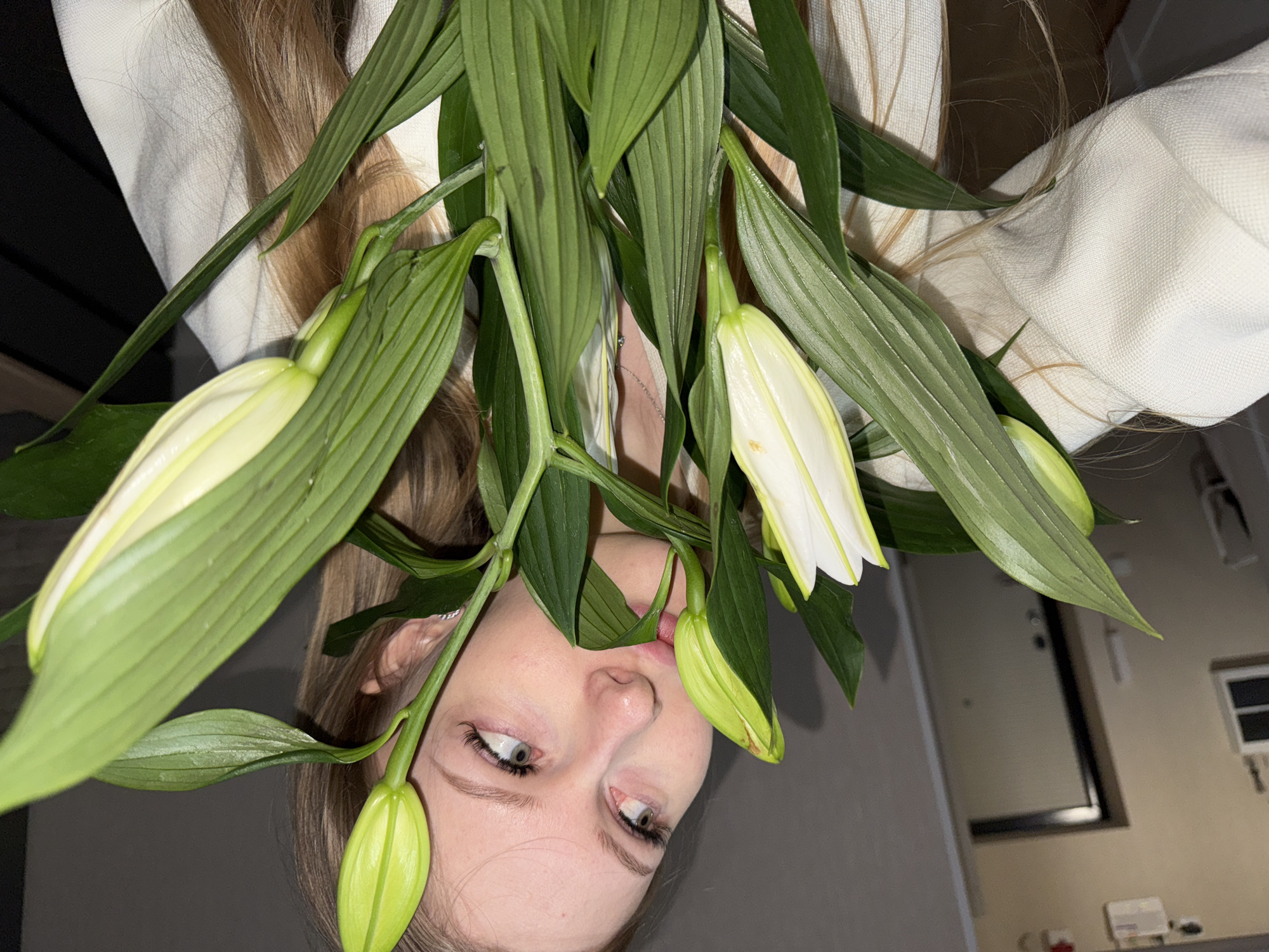 A young woman is positioned upside down in the frame, partially obscured by a bouquet of white and green lilies. Her face is visible beneath the blooms, with her eyes looking upward. She has blonde hair and is wearing a white garment. The background reveals a glimpse of an interior space, possibly a kitchen or dining area, with light-colored walls and a dark, rectangular object resembling a door or window frame. The location cannot be determined to be Perm, Russia from this image.