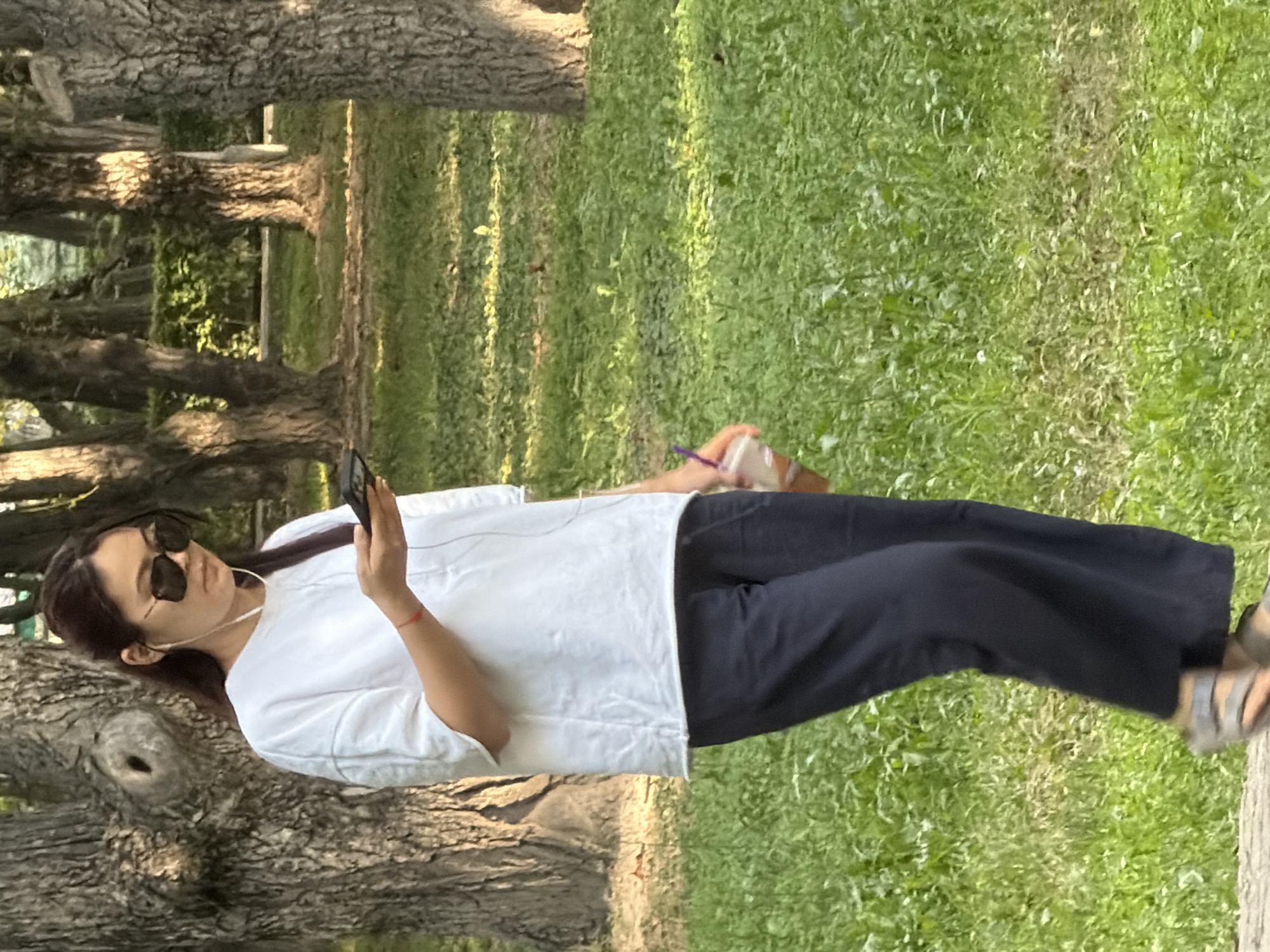 A medium-shot, full-body, outdoor, eye-level, natural light, portrait photo shows an East Asian woman walking in a park, possibly in a city. The woman is wearing a white t-shirt, dark blue pants, and sandals. She is also wearing black sunglasses and white earbuds, and holding a phone in her right hand. She has dark hair, pulled back from her face, and her left hand holds a plastic cup with a purple straw. The background consists of trees and grass. The woman's face is not clearly visible due to her sunglasses and the angle. The overall scene suggests a casual stroll in a green, urban environment.