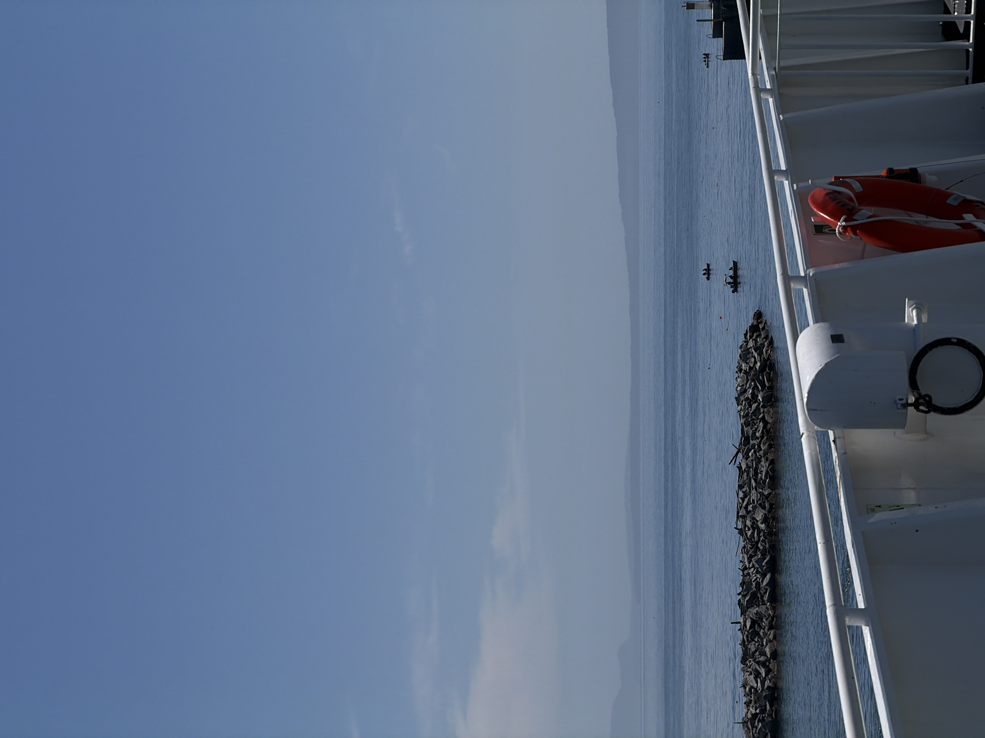 The image depicts a view from a ferry in Delta, Canada, looking out towards the water. In the foreground, the white railing of the ferry is visible, along with a red life preserver. Further out on the water, a rocky breakwater juts into the sea, and several small boats with people can be seen. In the distance, hazy mountains form the horizon under a clear blue sky.
