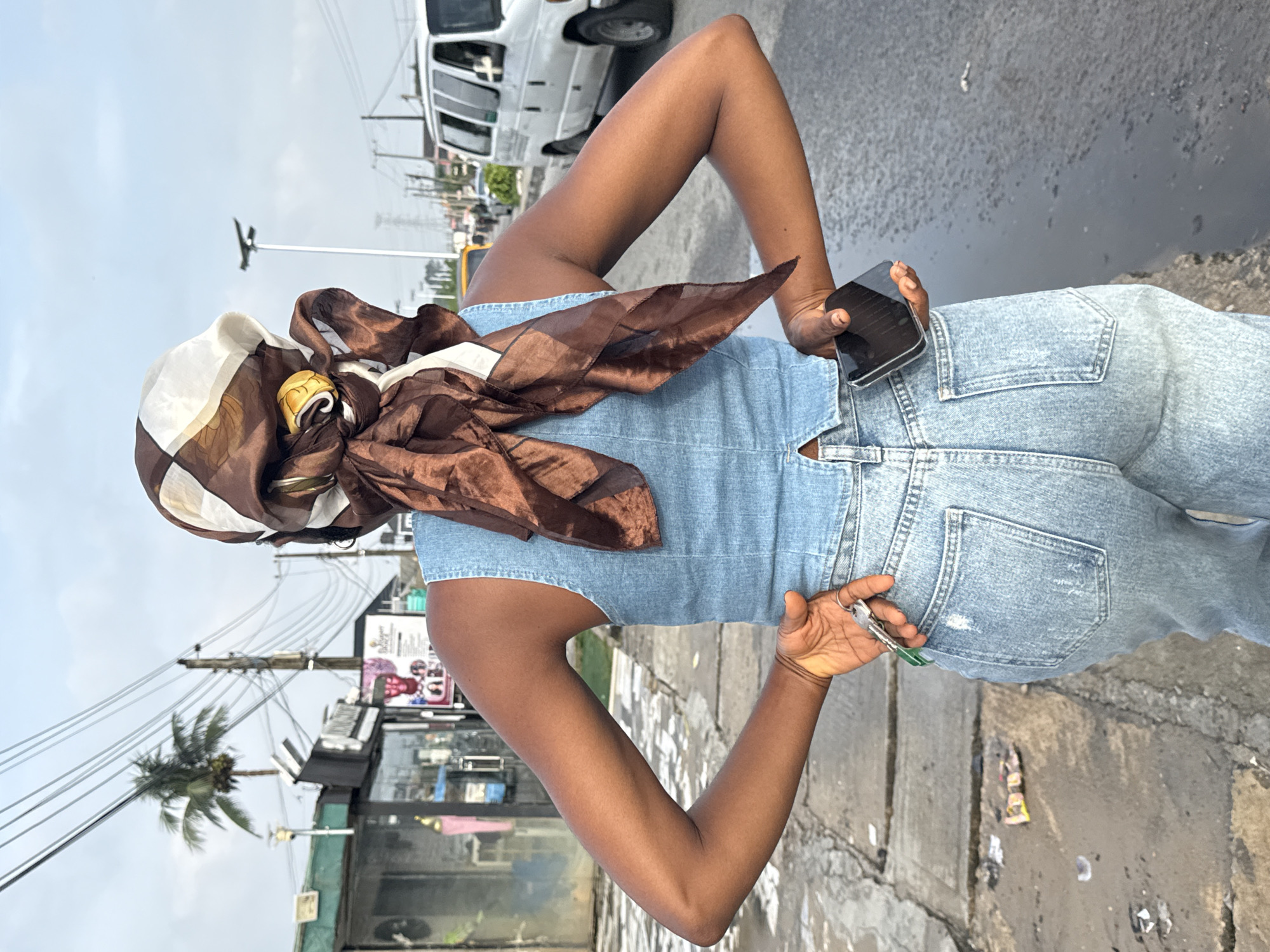 A person stands with their back to the camera, on a paved surface next to a street in Ado, Nigeria. They are wearing a blue denim sleeveless top and light blue distressed jeans. A brown and white patterned headscarf is tied around their head. Their right hand is on their hip, and their left hand holds a set of keys. A dark-colored smartphone is held in their right hand, tucked slightly behind them. In the background, there are utility poles with numerous wires, a palm tree, the side of a building with storefronts, and vehicles on the street including a white van and a yellow car. The sky is overcast. Debris is visible on the ground in the foreground.