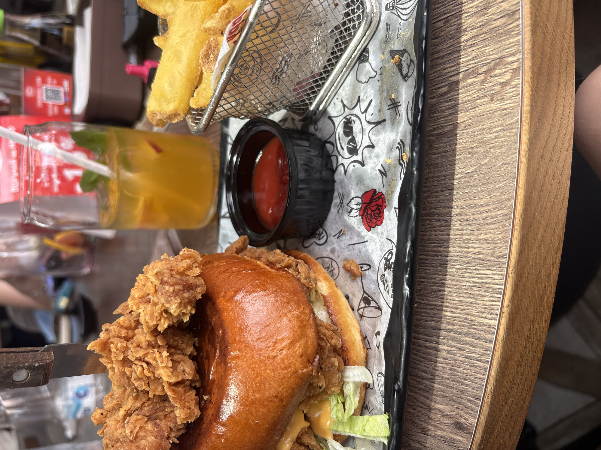This vertically oriented, brightly lit image captures a casual meal set on a wooden table in what appears to be a restaurant or cafe, likely in Sofia, Bulgaria given the provided context. The main subjects are a substantial fried chicken sandwich, a side of fries, ketchup, and a drink.
The fried chicken sandwich is prominently featured, boasting a shiny, golden-brown brioche bun, a large, crispy piece of fried chicken, fresh shredded lettuce, and a creamy, light orange sauce. A knife with a rustic wooden handle is visible near the sandwich.
Next to the sandwich, a small metal basket holds a generous serving of thick-cut, golden-brown fries. A black plastic cup filled with vibrant red ketchup is placed beside the fries.
The accompanying beverage, served in a glass jar, is a refreshing yellow-orange drink, garnished with green mint leaves and a hint of red fruit, complete with a white straw.
The food items rest on a black serving tray lined with white paper. This paper features an array of stylized black and white cartoon-like illustrations, including a rose, a bomb, a speech bubble, and a symbol resembling a bat, with a partially visible text that appears to be "Burg...". The wooden table beneath has a distinct grain pattern. In the blurred background, a red sign with a QR code and unreadable text can be discerned, along with a partial view of a person's arm or leg to the far right, indicating someone is seated at the table. The overall scene suggests an appetizing daytime meal.
