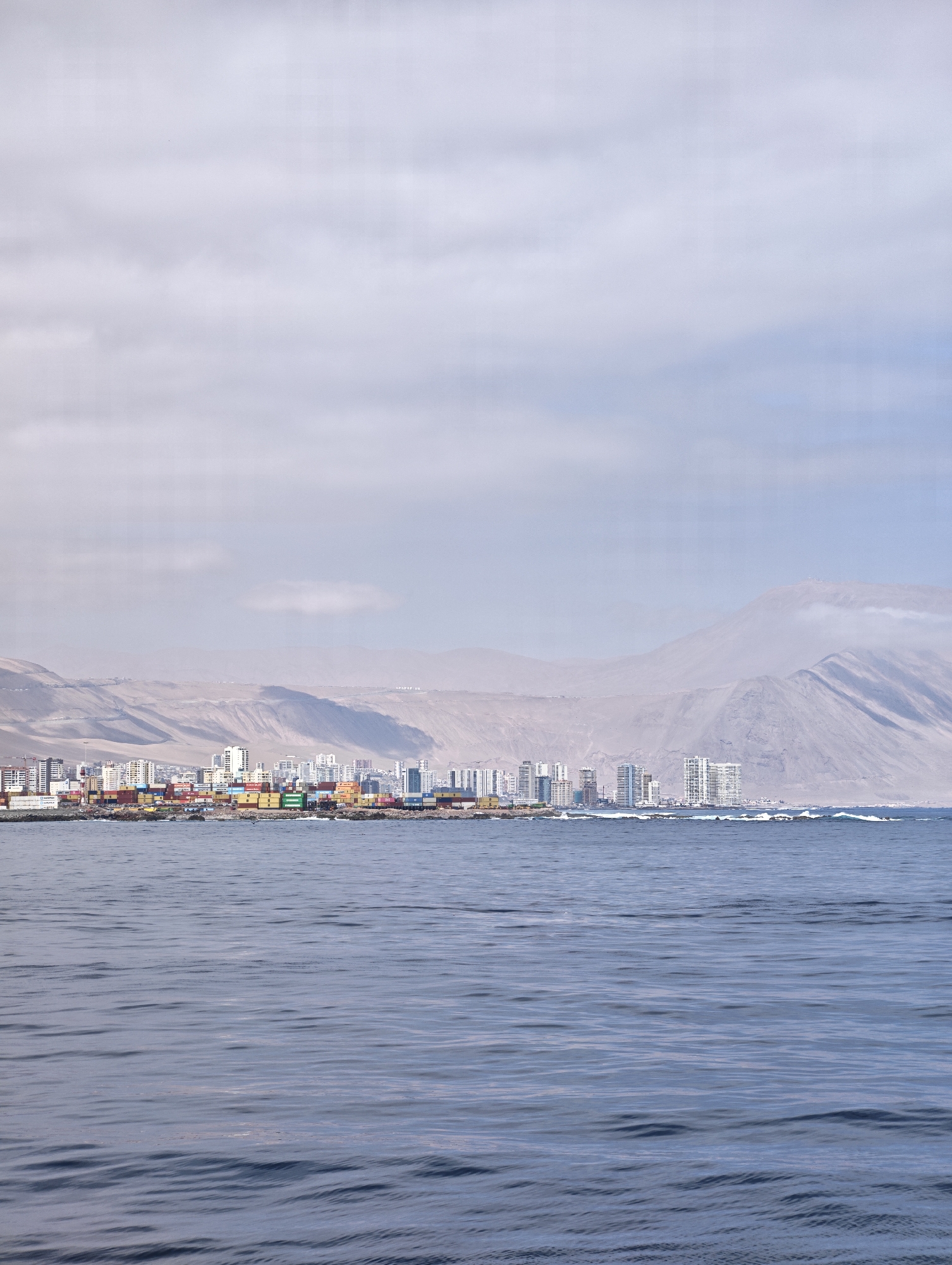 The image shows a panoramic view of the city of Iquique, Chile, from the ocean. In the foreground, the deep blue water of the Pacific Ocean is calm with gentle ripples. The middle ground features the coastline of Iquique, with a dense cluster of buildings, including high-rise apartments and office buildings, lining the shore. Several shipping containers are visible stacked on the docks, indicating the presence of a port. Behind the city, arid, sandy hills rise up, characteristic of the Atacama Desert region. The sky above is overcast with a pale, hazy light. The scene captures the urban landscape of Iquique juxtaposed with its natural desert environment and the vast ocean.