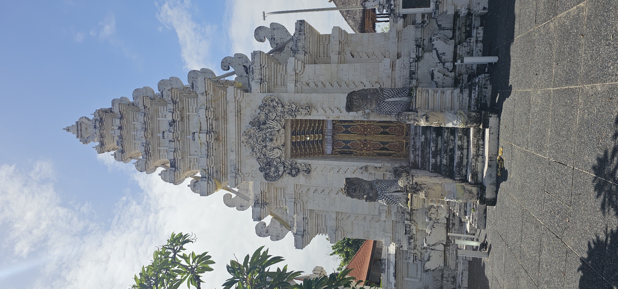 The image presents a view of a traditional Balinese temple structure in Kuta Selatan, Indonesia. The temple is constructed primarily of a light-colored stone, possibly limestone or a similar material, with intricate carvings and tiered rooflines characteristic of Balinese architecture. The structure features ornate gates or doorways decorated with elaborate gold-colored details, flanked by guardian statues clad in checkered textiles. The foreground includes paving stones, suggesting a pathway leading to the temple entrance, and foliage from nearby trees is visible at the bottom of the frame. The sky is blue with scattered clouds, indicating a clear day.