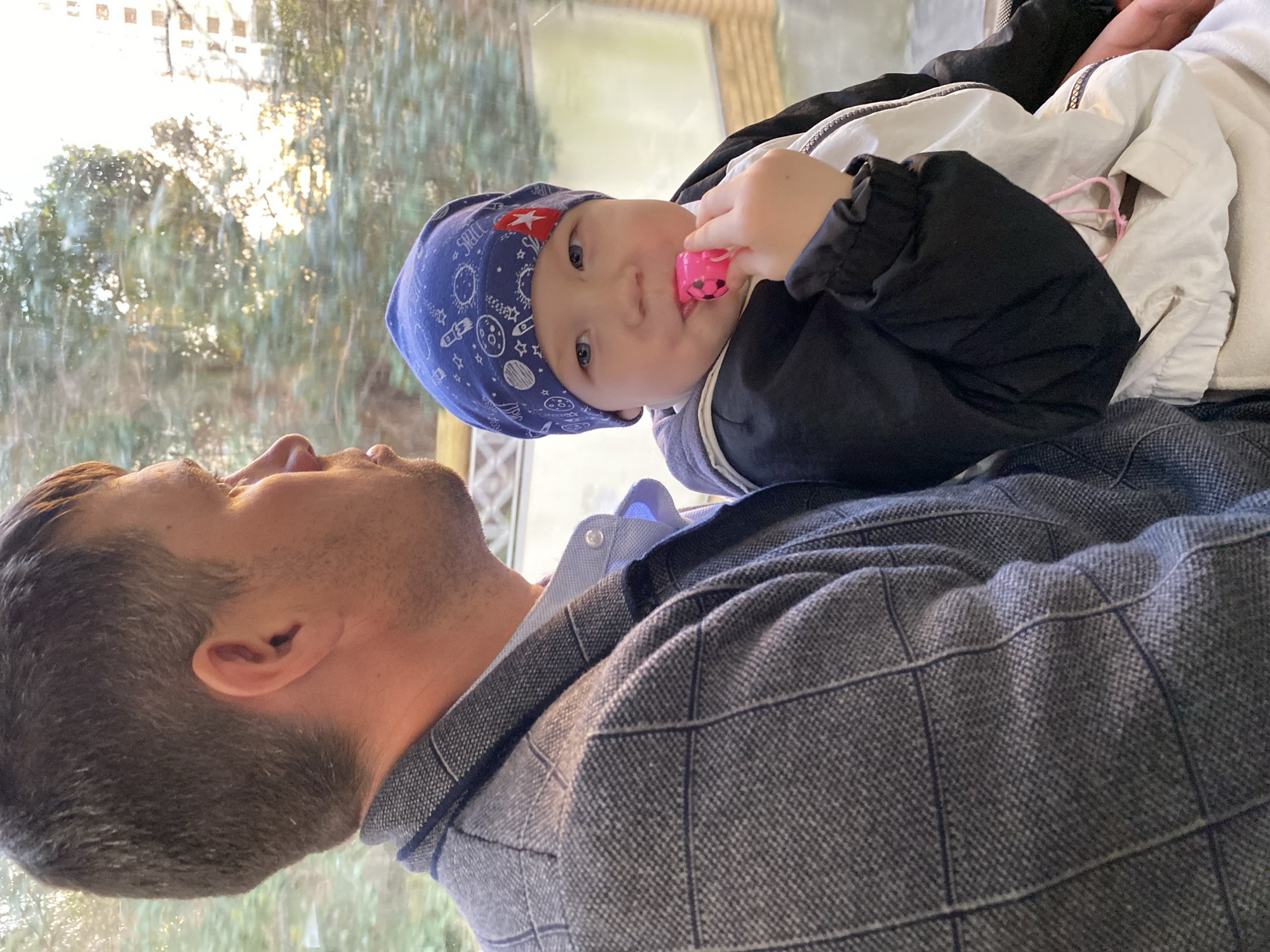 A baby in a blue hat with a red star sits on the chest of a man. The baby is fair-skinned, has blue eyes, and is holding a pink toy. The man has short brown hair and a short beard, and is wearing a gray patterned shirt. Outside the window is a view of trees. The location is Sochi, Russia.