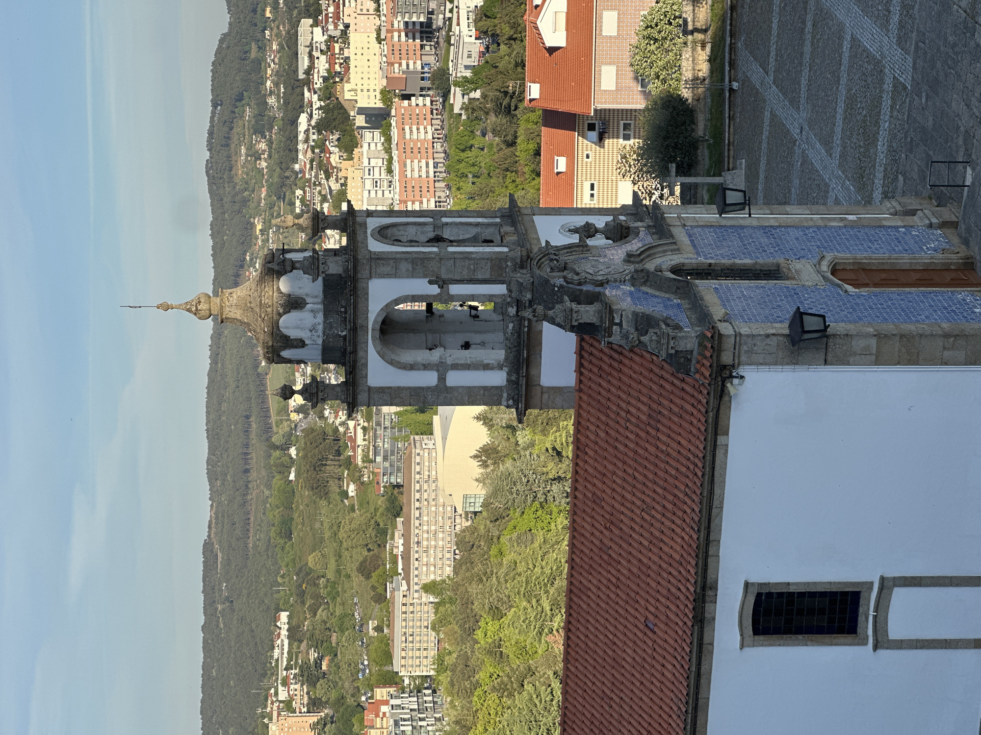 An aerial view depicts a church tower, potentially Igreja de São Pedro Vila Real in Vila Real, Portugal. The tower's architectural style suggests a baroque or similar historical influence, with a dome-like structure atop the tower. The tower has a white facade with a red-tiled roof. In the background are buildings and greenery, indicating an urban landscape set against a natural backdrop. The sky is clear, suggesting good weather conditions.