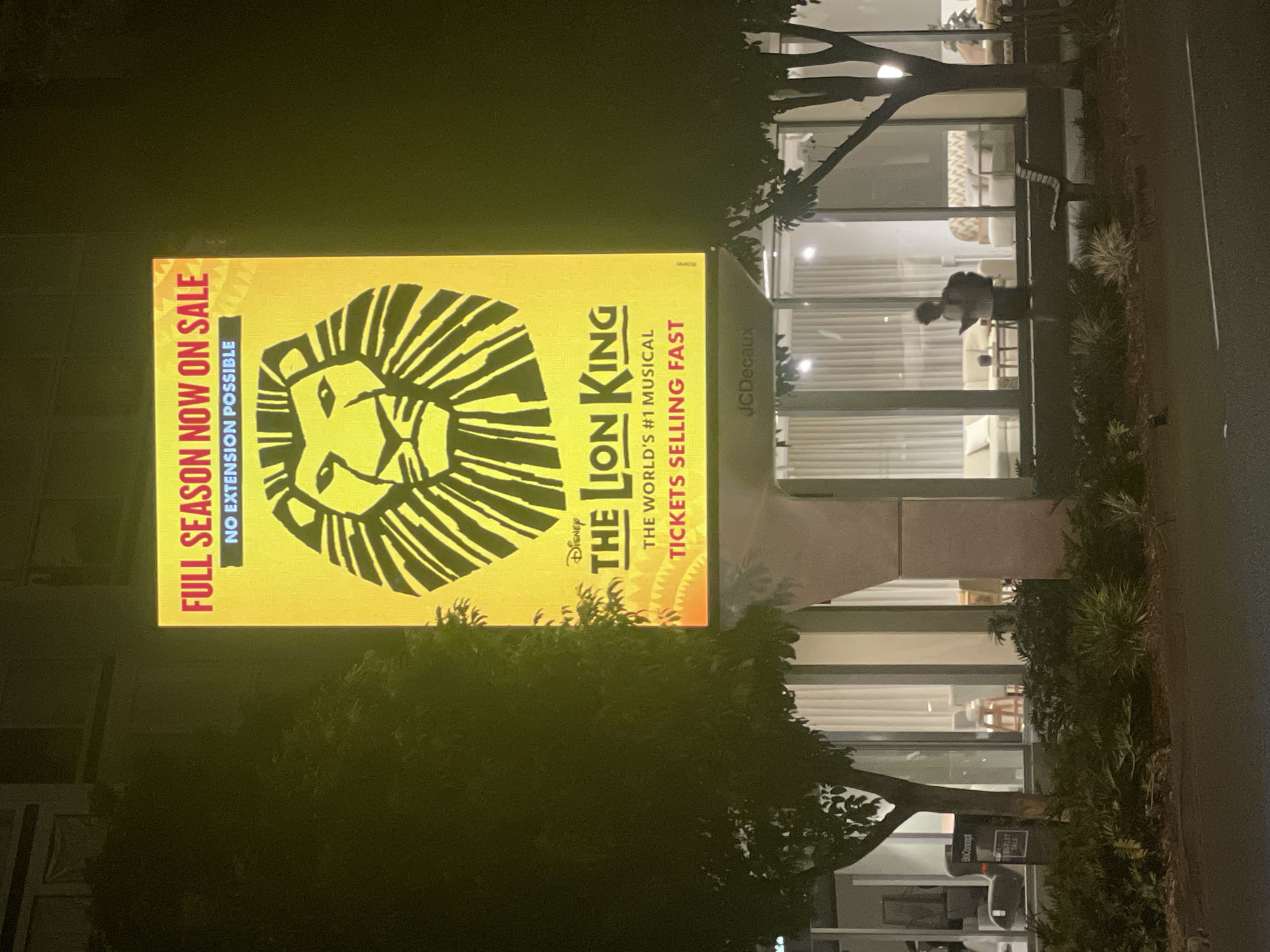 The image shows a large, illuminated billboard for Disney's "The Lion King" musical. The billboard features the iconic lion's head logo and text that reads "FULL SEASON NOW ON SALE NO EXTENSION POSSIBLE THE WORLD'S #1 MUSICAL TICKETS SELLING FAST." The billboard is positioned above a building with large windows, displaying indoor furniture. To the left of the billboard is a tree, and to the right is another tree with a silhouette of a person standing in front of the building. The scene appears to be at night, as indicated by the lighting and the dark environment. Based on the presence of the JCDecaux advertisement panel on the lower part of the billboard structure, this scene is likely located in a public urban space. The architecture of the building and the type of billboard suggest a commercial or entertainment district. Without further contextual information or specific landmarks visible, pinpointing the exact location within Brisbane, Australia is not possible from the image alone.